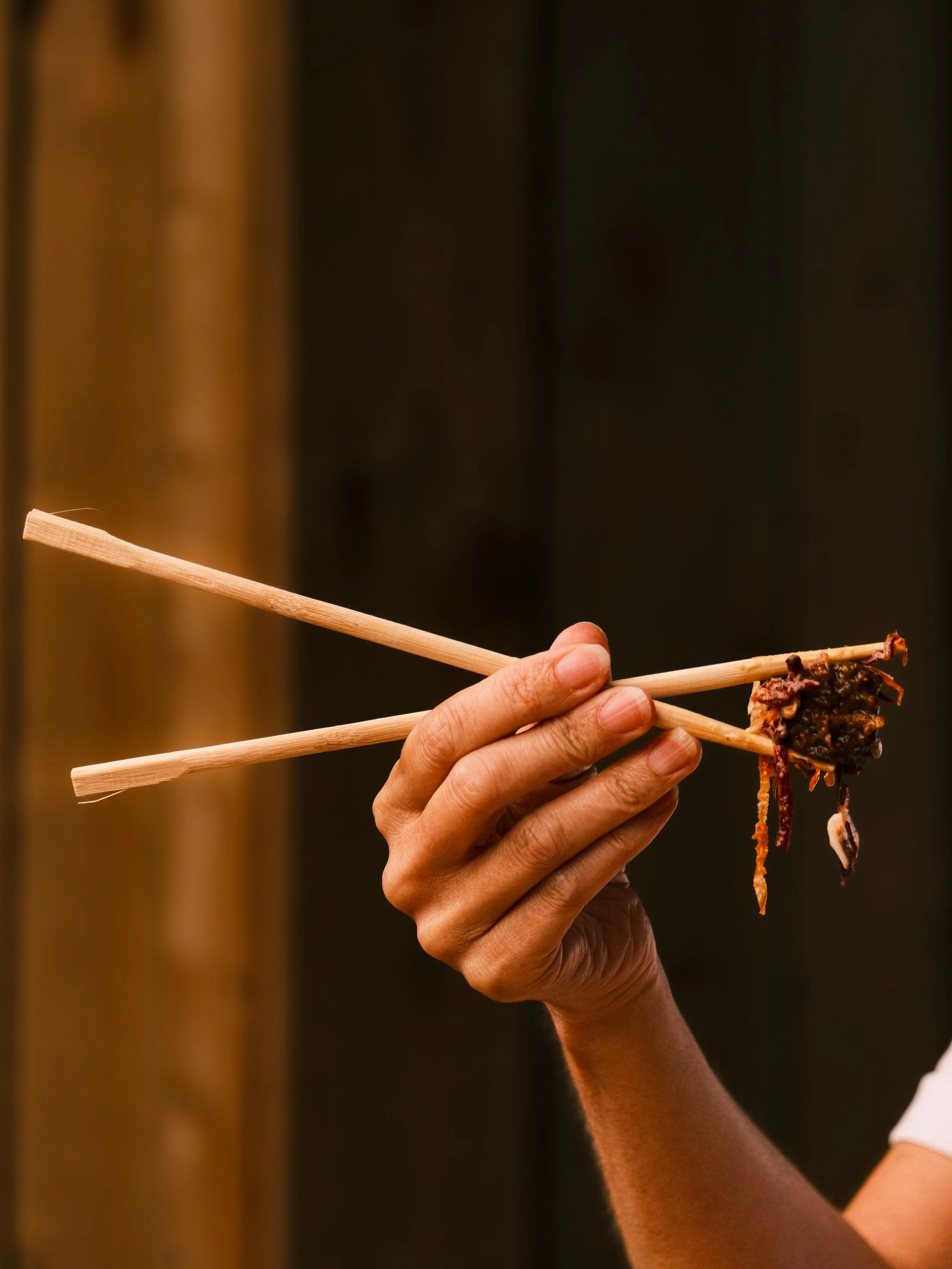 A person's hand holds a pair of chopsticks with a piece of cooked meat against a dark background.