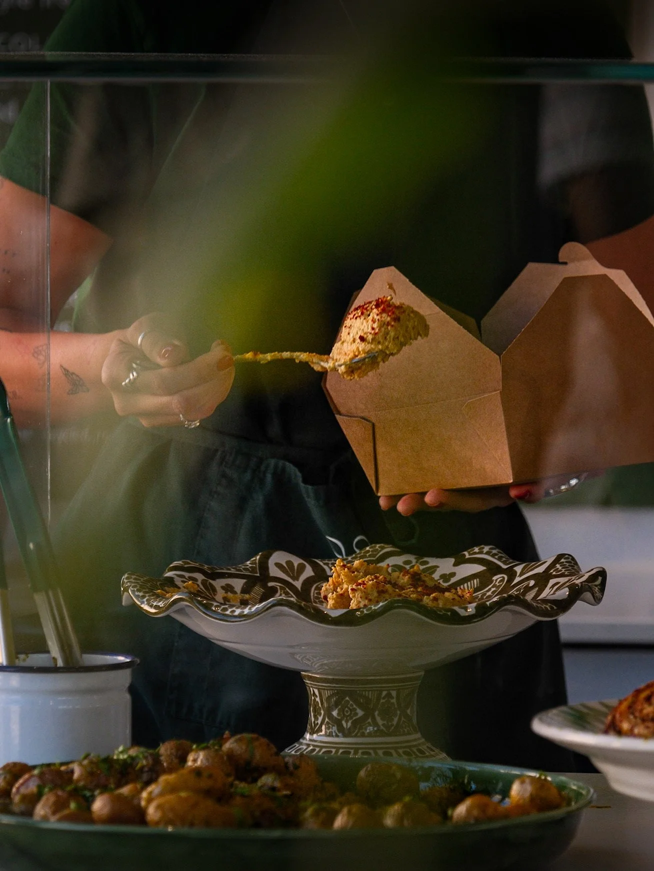 Person holding a cardboard takeout box with a slice of cake inside, standing behind a decorative cake stand with baked goods, in a display case.