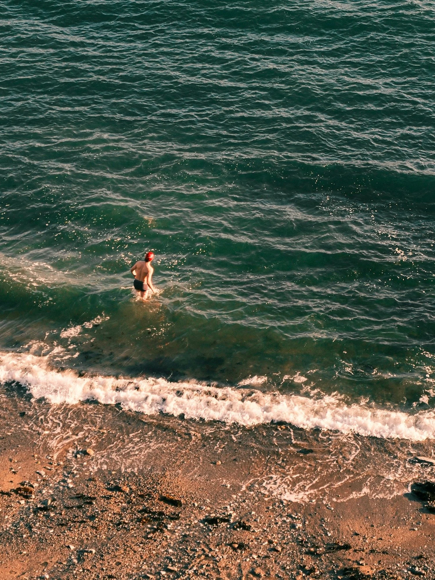 A person in a red swim cap and black swim trunks standing in the ocean near the shoreline, with small waves and pebbles on the beach.