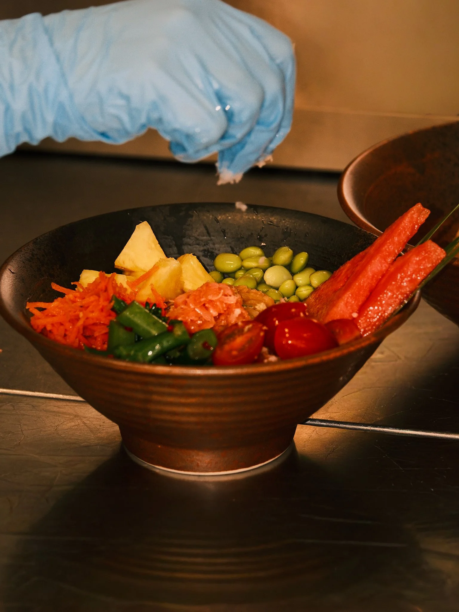 Close-up of a bowl with various colorful Asian dishes, with a hand wearing a blue glove sprinkling something into the bowl.