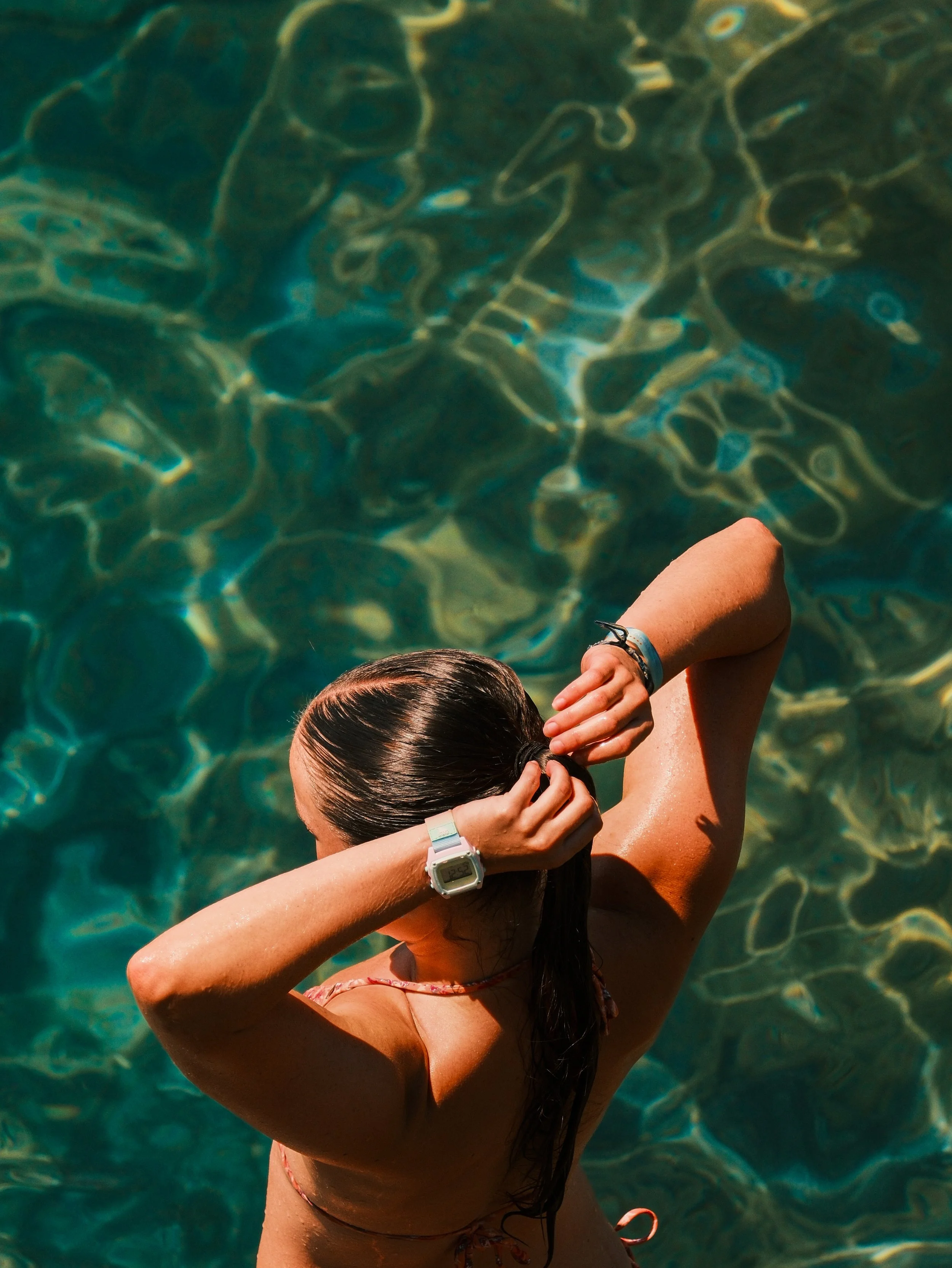 A woman with wet hair taking a selfie from above while in a swimming pool, with her arms raised and holding her hair back.