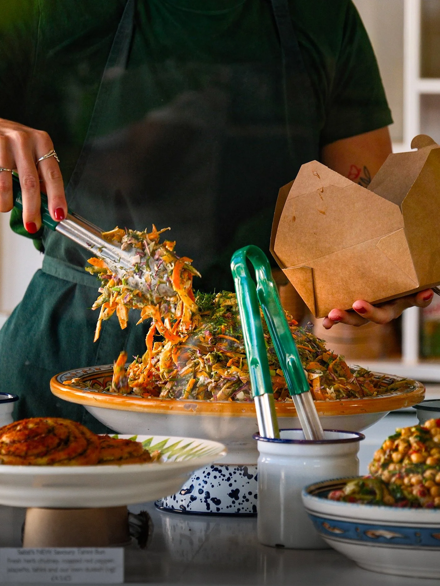 Person serving a large salad from a dish onto a plate, with other dishes of food on the table.