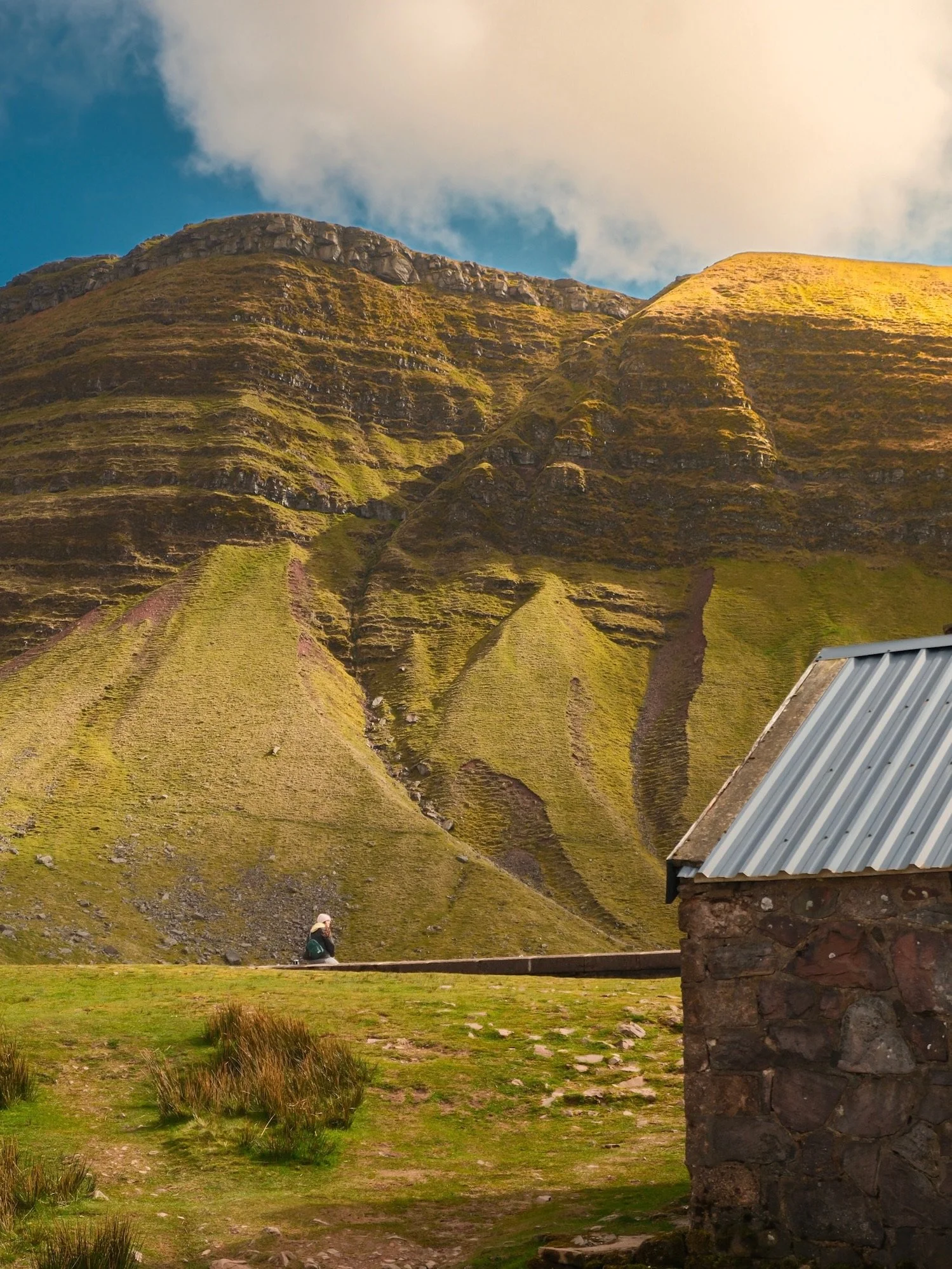 A person with a hat and backpack sitting on a bench in front of a green hillside with an ancient stone building with a metal roof in the foreground, under a partly cloudy sky.