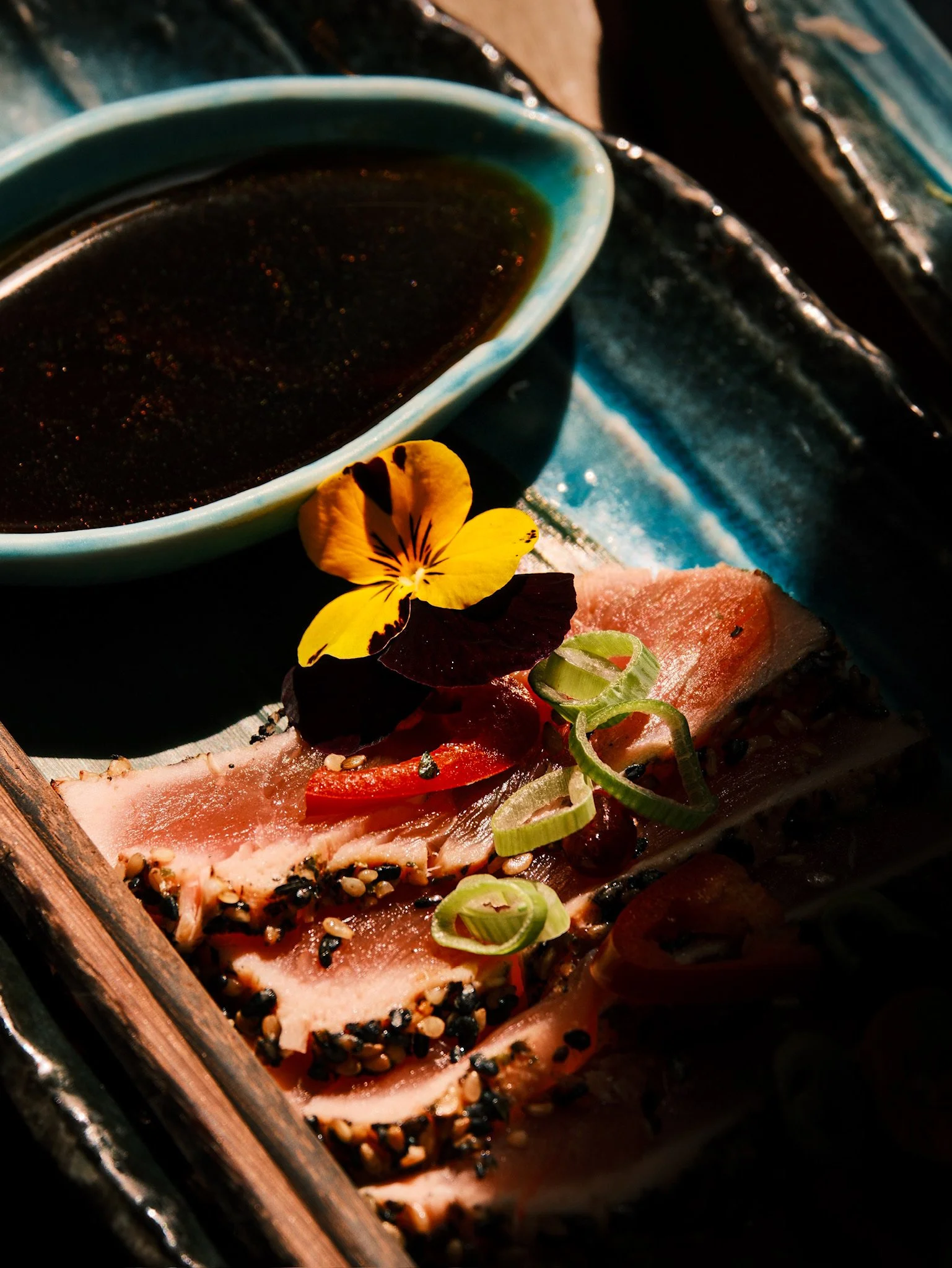 Close-up of a sushi platter with slices of fish topped with sesame seeds, garnished with sliced green onions, a yellow edible flower, and a side of soy sauce in a green bowl.
