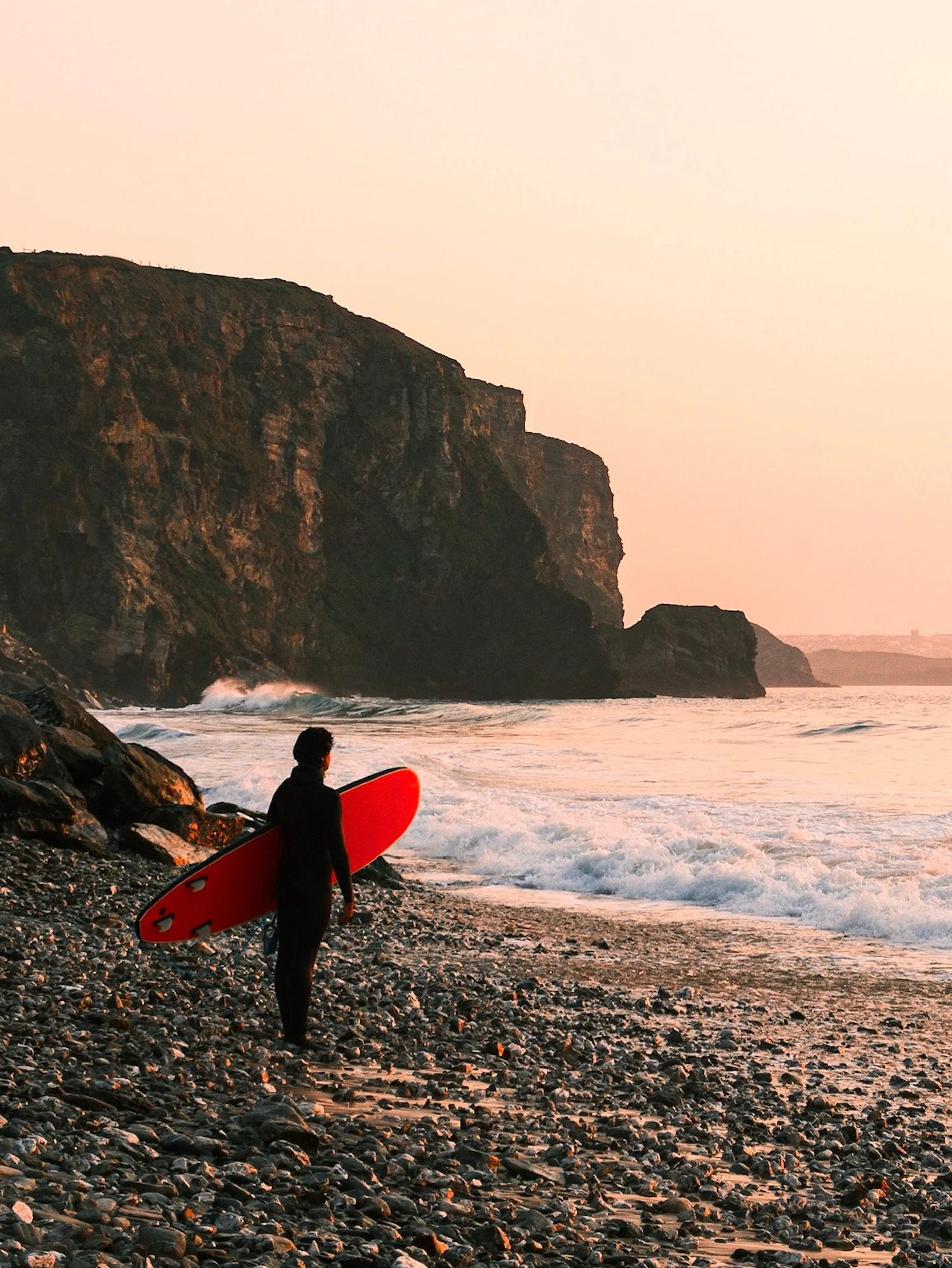 A person holding a red surfboard on a rocky beach, with cliffs to the left and ocean waves, during sunset or sunrise.