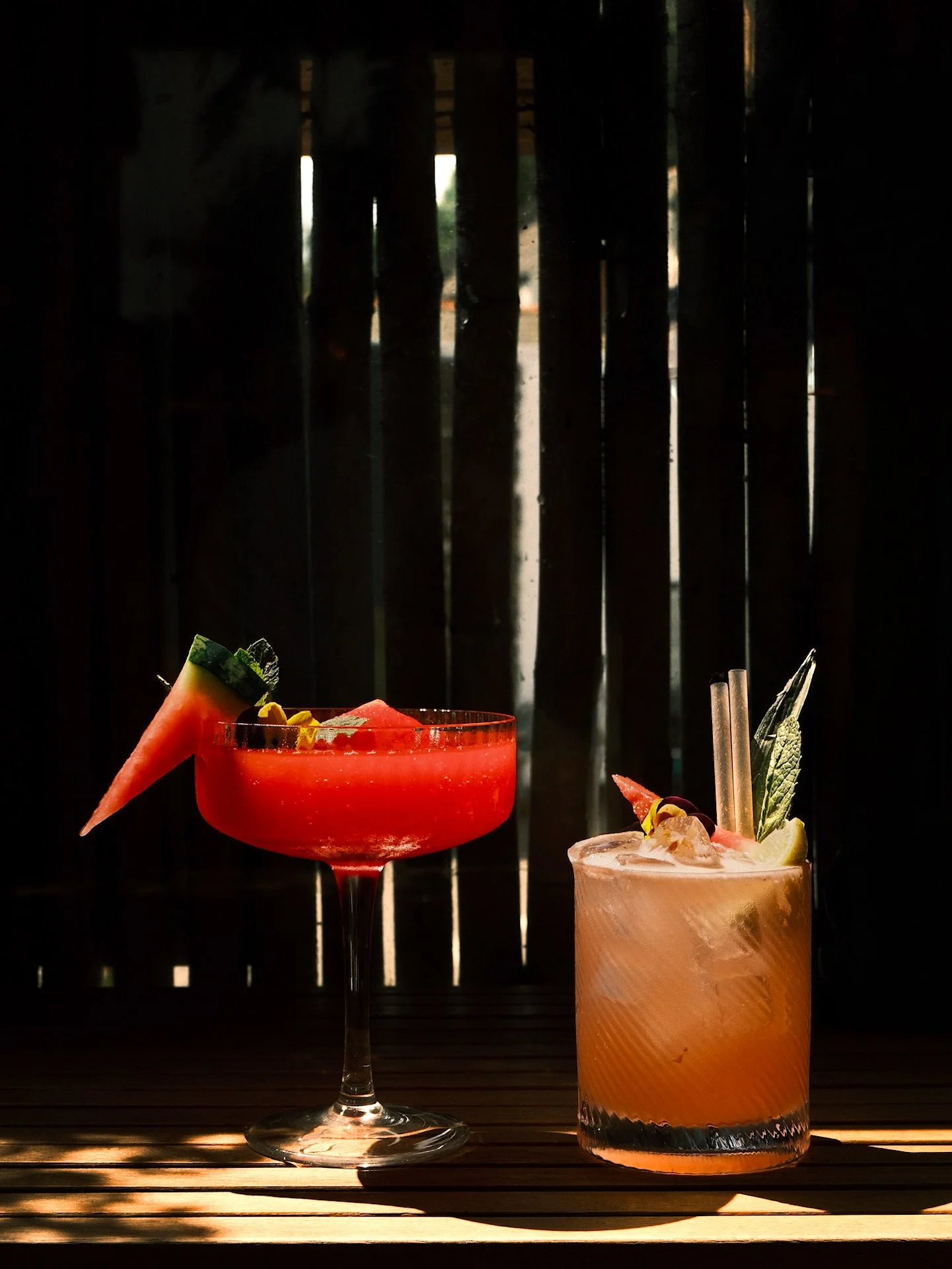 Two colorful cocktails on a wooden surface with a dark background and vertical slats, garnished with fruit and herbs.