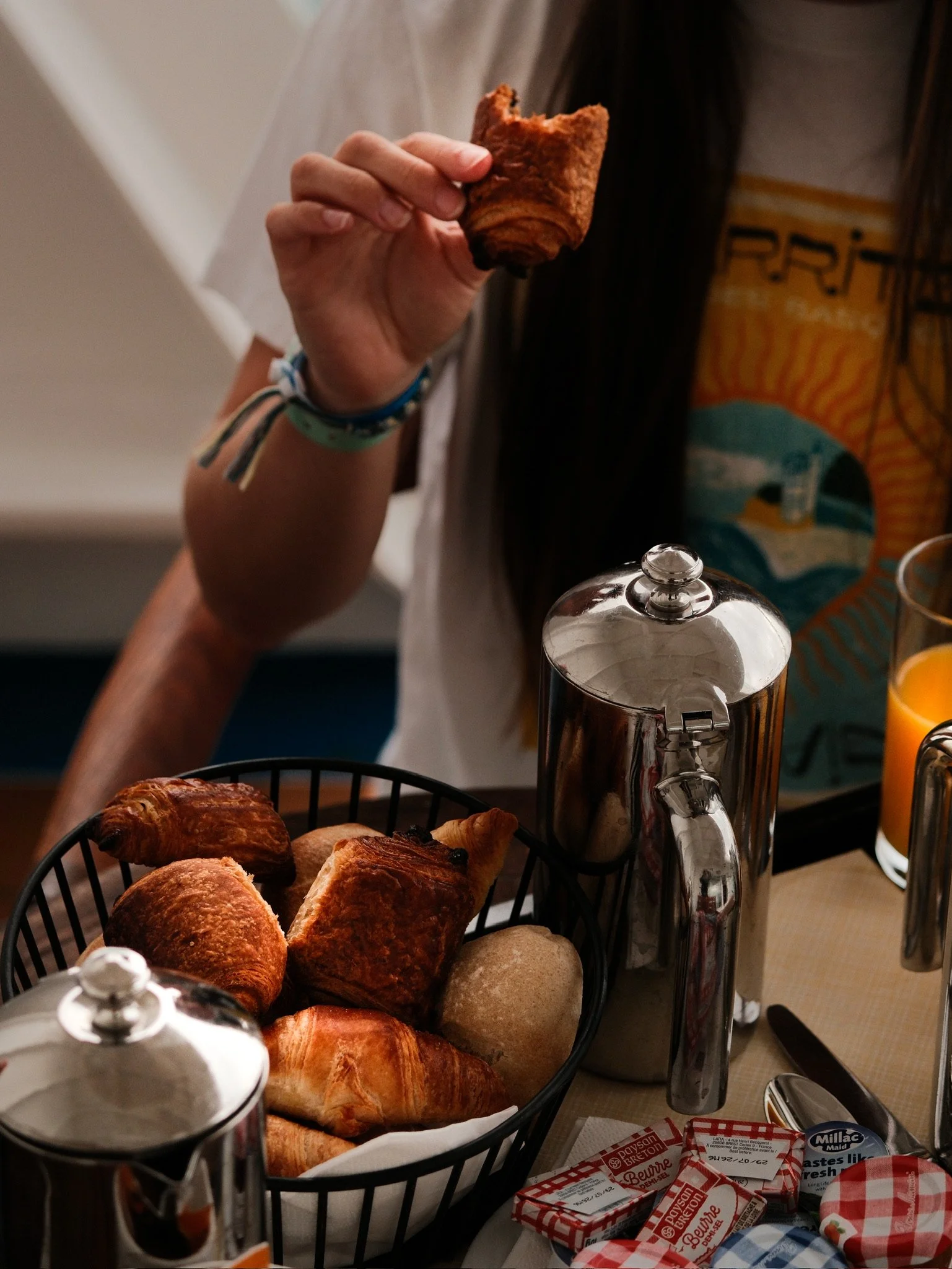 A person holding a croissant above a basket of assorted baked goods, with butter and jam packets nearby on a table.