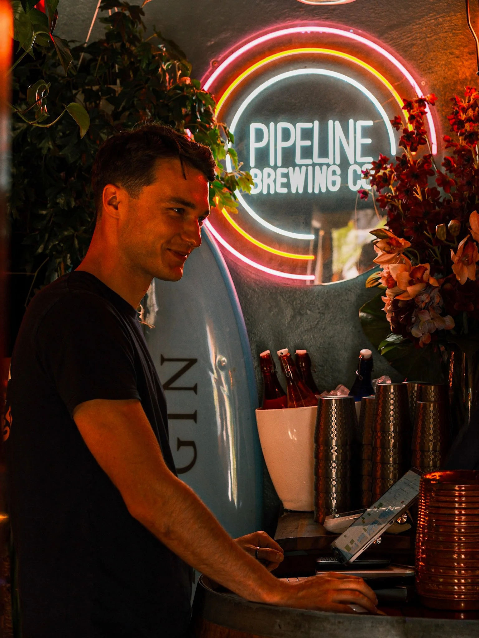 Man preparing drinks at a bar counter under a neon sign that reads 'Pipeline Brewing Co', surrounded by plants and stacked cups.