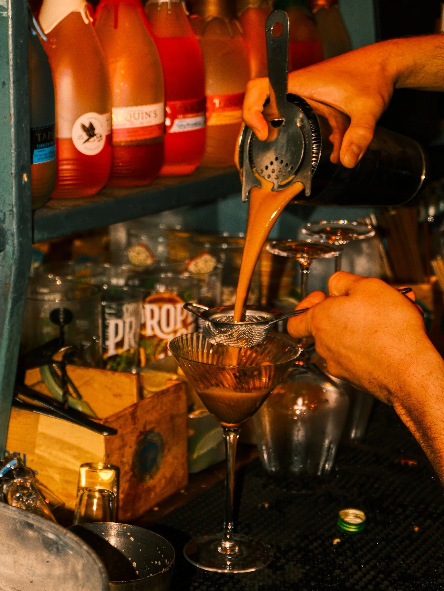 A bartender pouring a brown liquid, likely a cocktail, through a strainer into a cocktail glass with a long stem and wide bowl.
