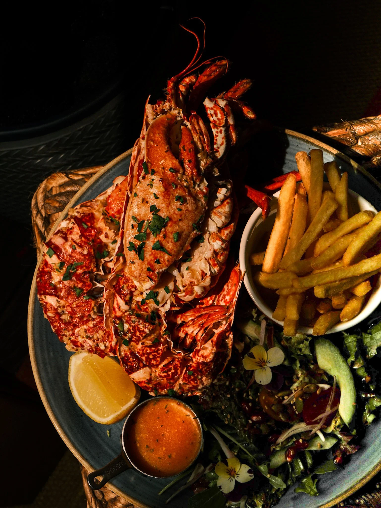 Lobster served on a plate with lemon wedges, fries, and salad, accompanied by two dipping sauces.