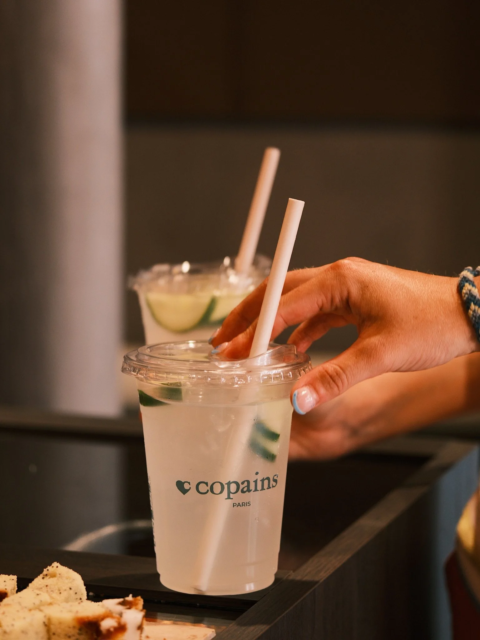 A hand holding a plastic cup with a drink that has lime slices and two white straws, with a dark background and some cake slices in the foreground.