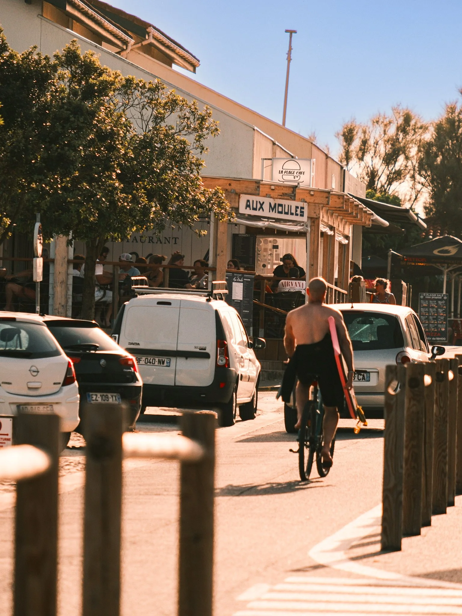 A person riding a bicycle along a sidewalk in a parking lot, with cars parked nearby and a building with signage reading 'AUX MOULES' and 'LA PLAGE CAFE' in the background.