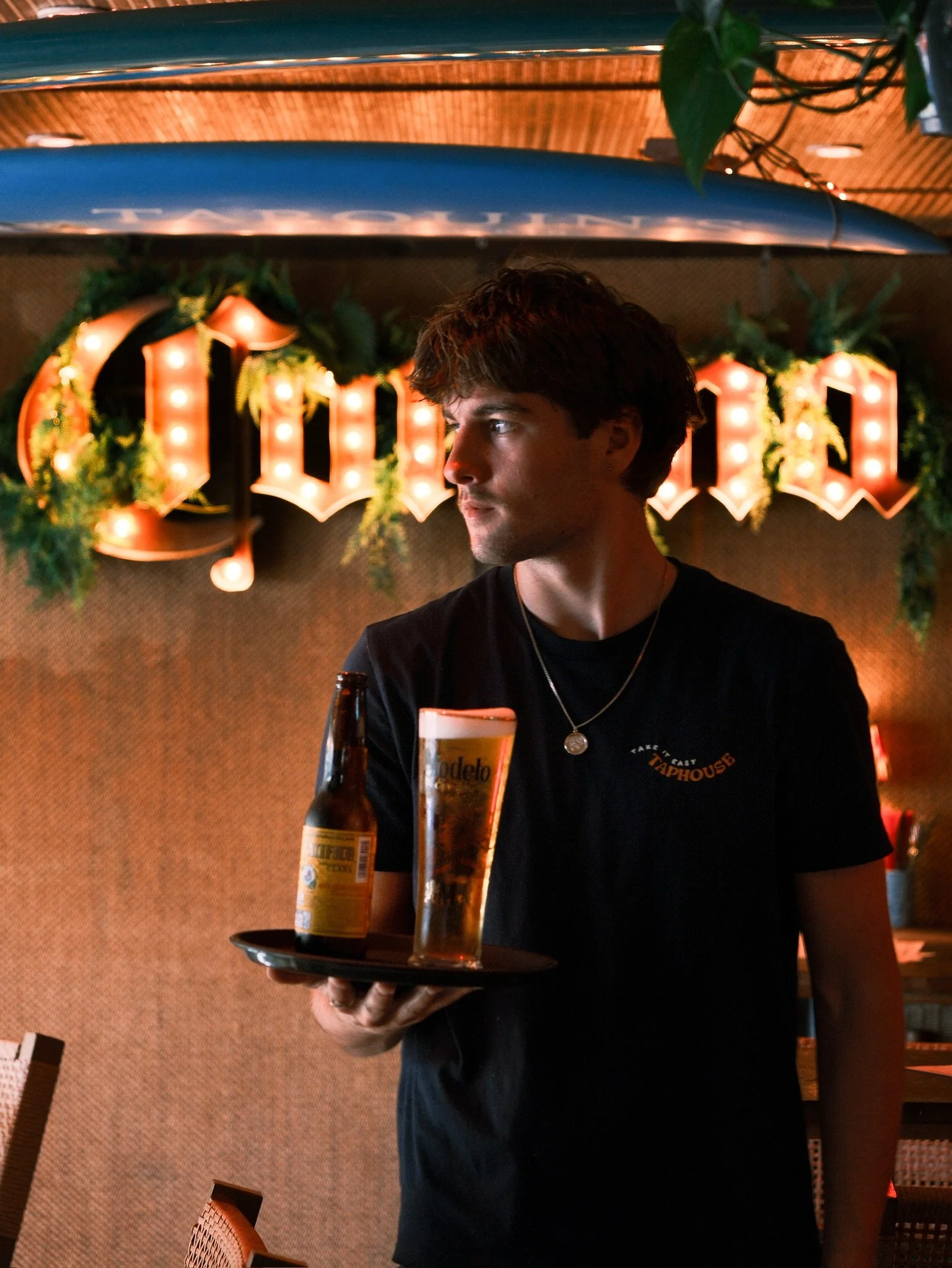 A person with dark hair wearing a black T-shirt holding a tray with a beer bottle and a glass of beer in a dimly lit restaurant or bar. There is a large illuminated sign in the background.
