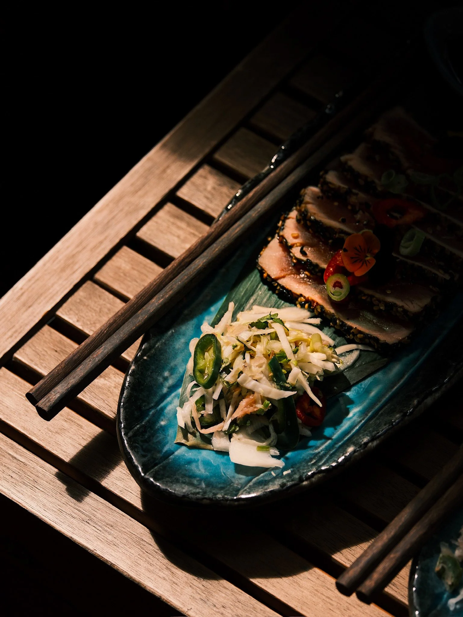 Close-up of a blue ceramic plate with sliced cooked fish topped with sesame seeds, garnished with edible flowers, and accompanied by a side of shredded cabbage salad with sliced green chili, all on a wooden table.