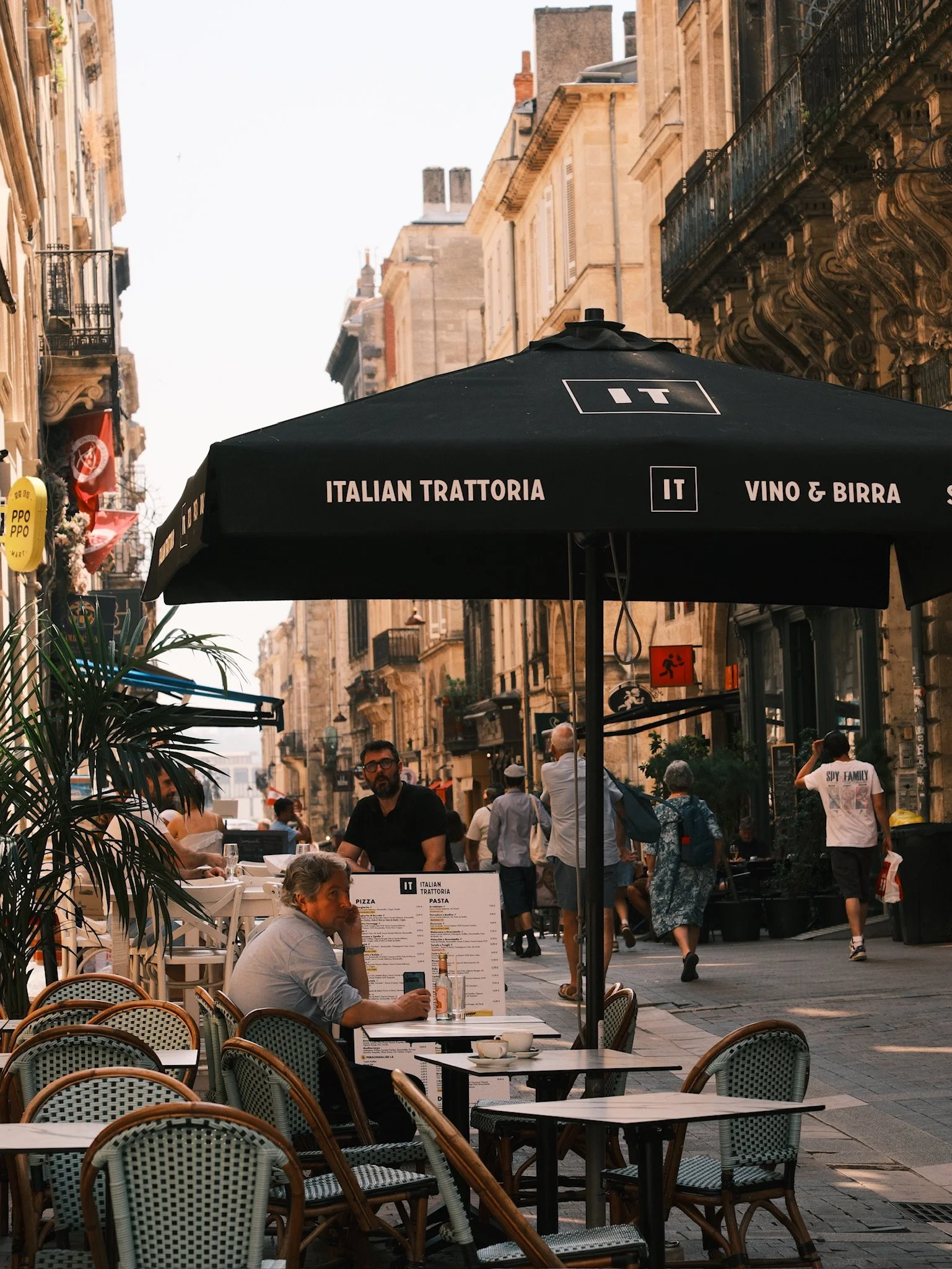 Outdoor seating area of an Italian restaurant with a black umbrella that says "Italian Trattoria" and "Vino & Birra," with people dining and walking on a European city street lined with historic buildings.