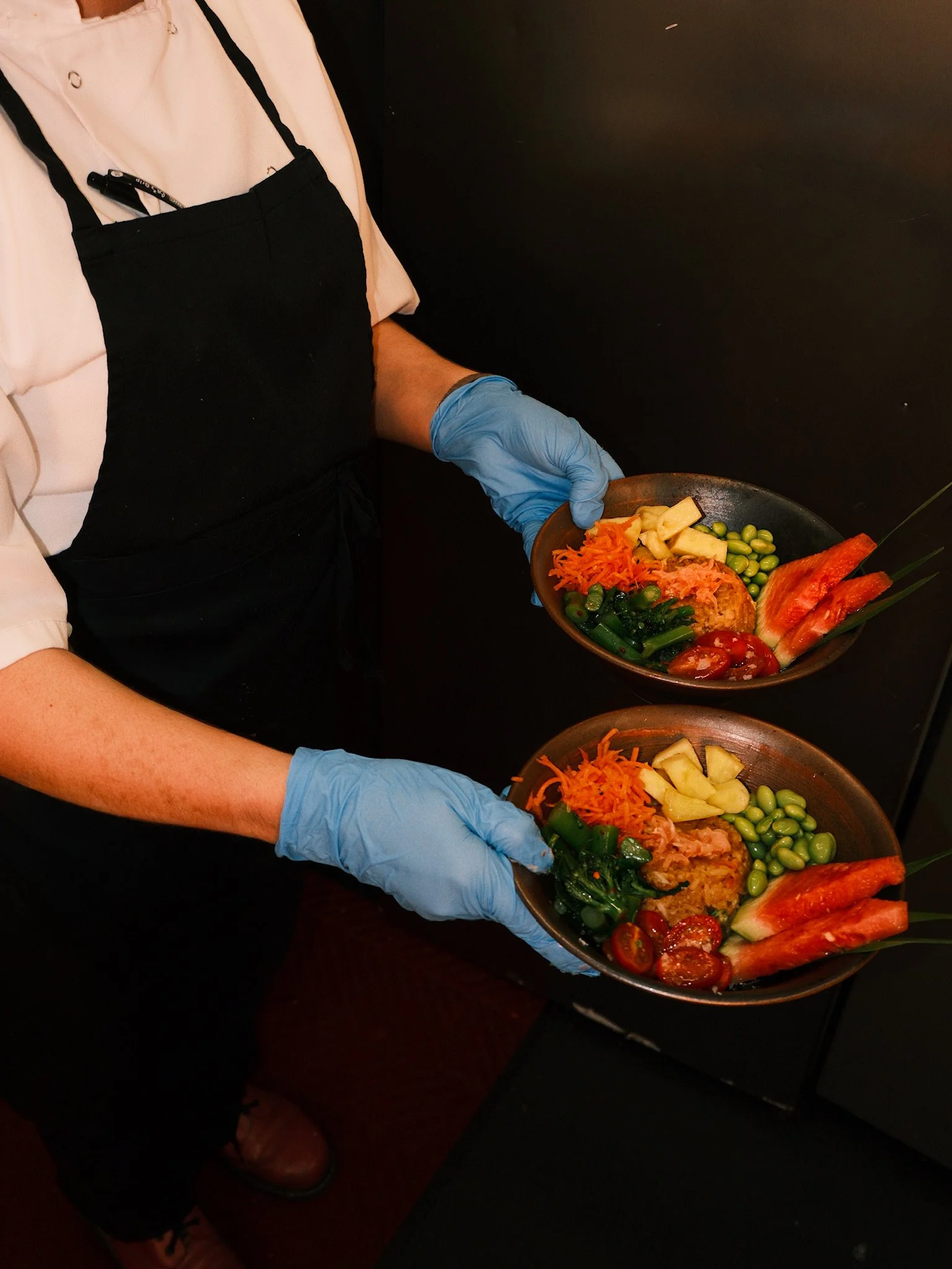 A person wearing a white shirt and black apron with blue gloves holding a bowl of colorful chopped vegetables and fruits, including carrots, edamame, pineapple, cherry tomatoes, and watermelon slices.