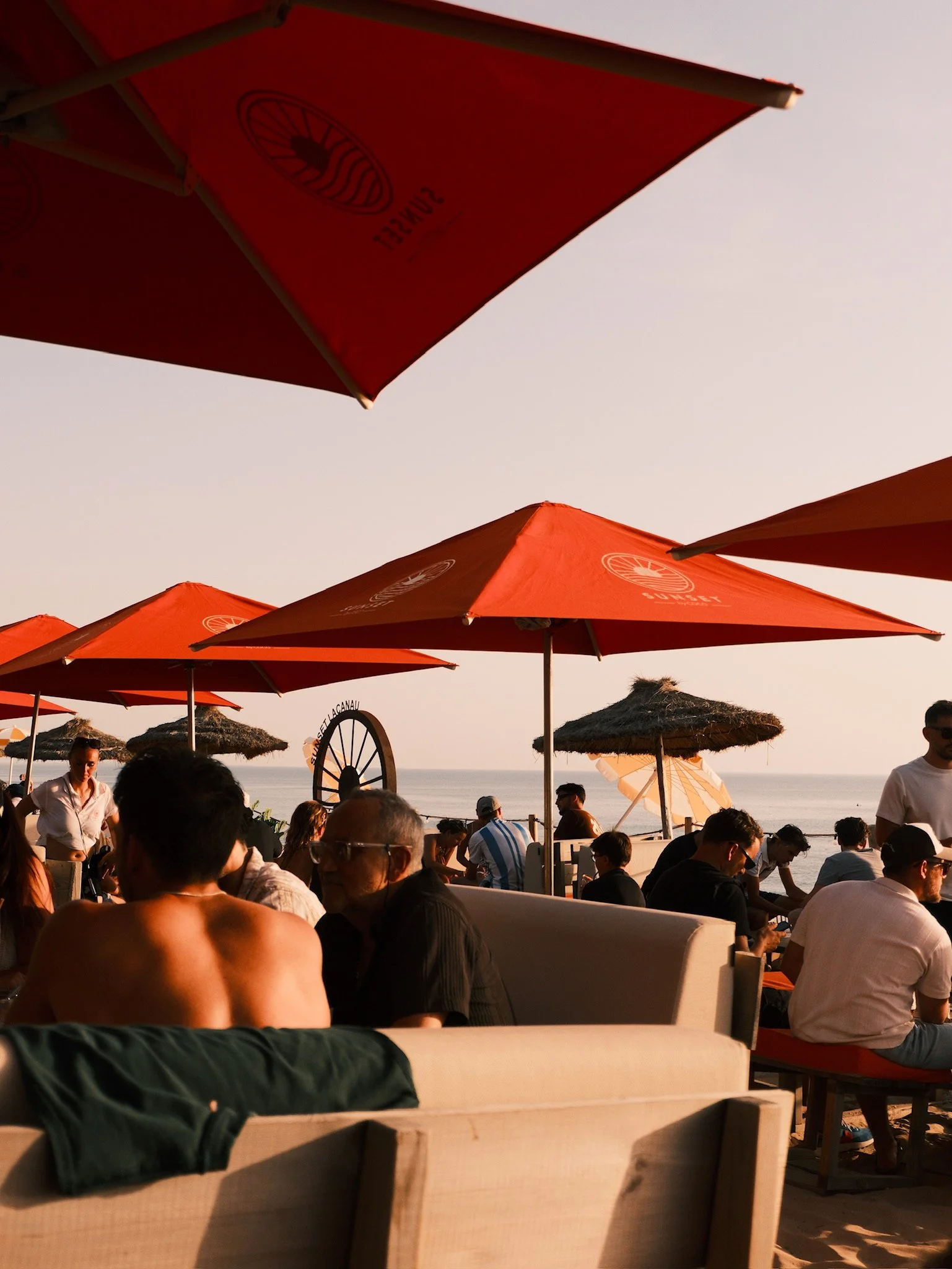 Beachside scene with many people sitting under red umbrellas, some in sun hats, and thatched-roof cabanas along the shoreline, with the ocean and sky in the background during sunset.
