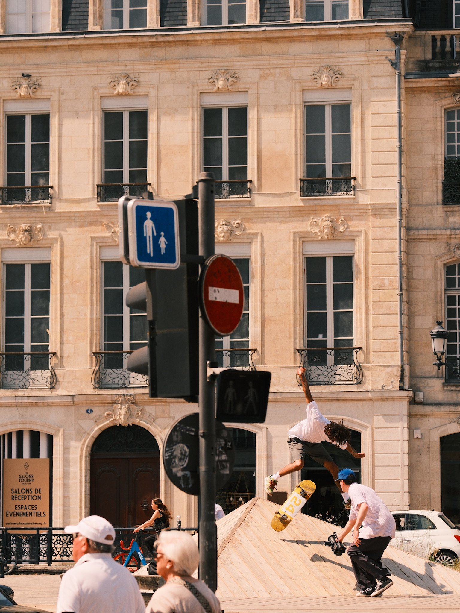 A skateboarder is mid-air performing a trick off a ramp in an urban area, with several onlookers and a historic building in the background.