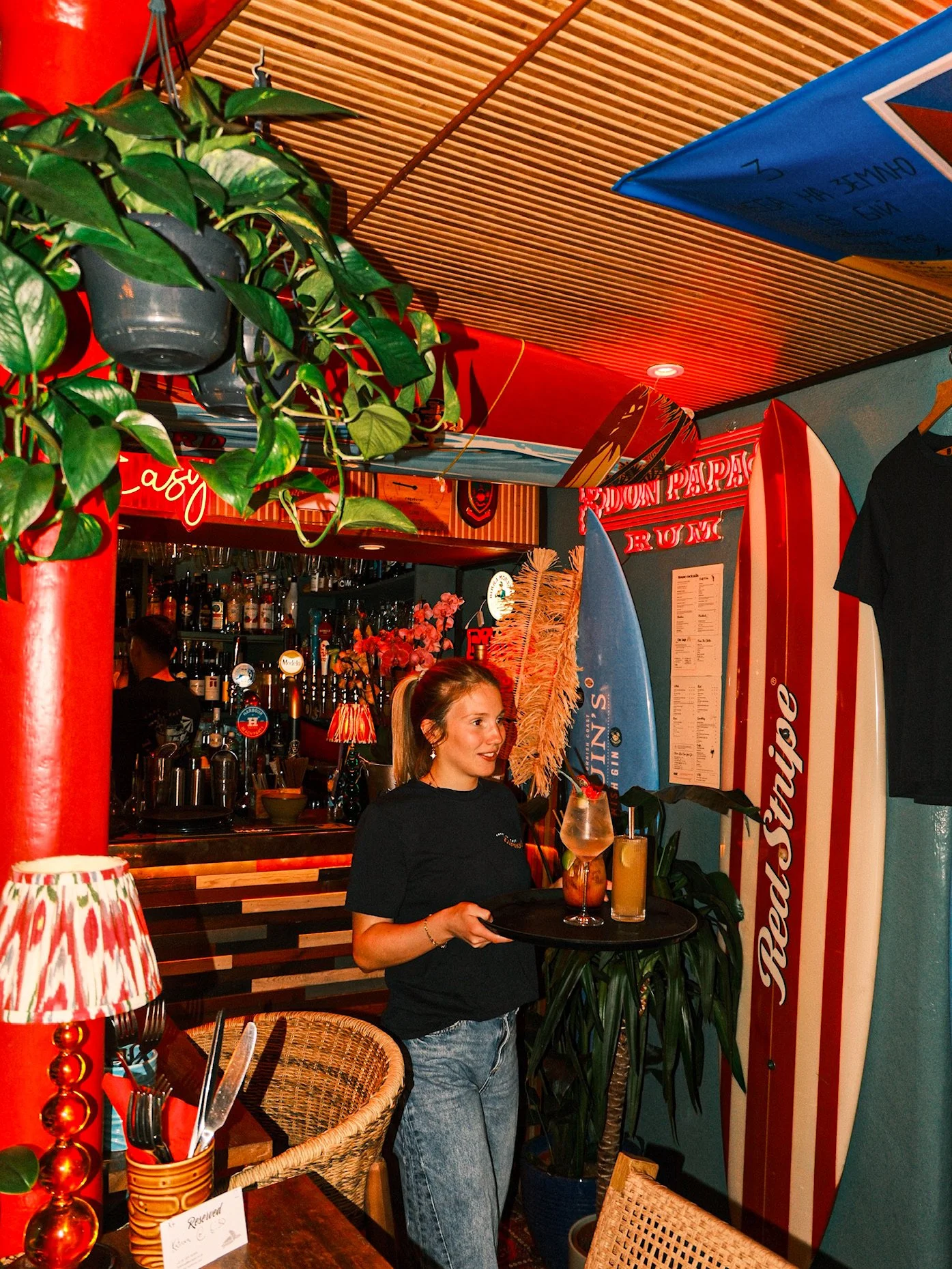 A young woman with long hair wearing a black T-shirt and jeans is holding a tray with bar drinks in a colorful, tropical-themed bar or restaurant. The bar has surfboards, neon signs, and vibrant decor elements, creating a lively atmosphere.