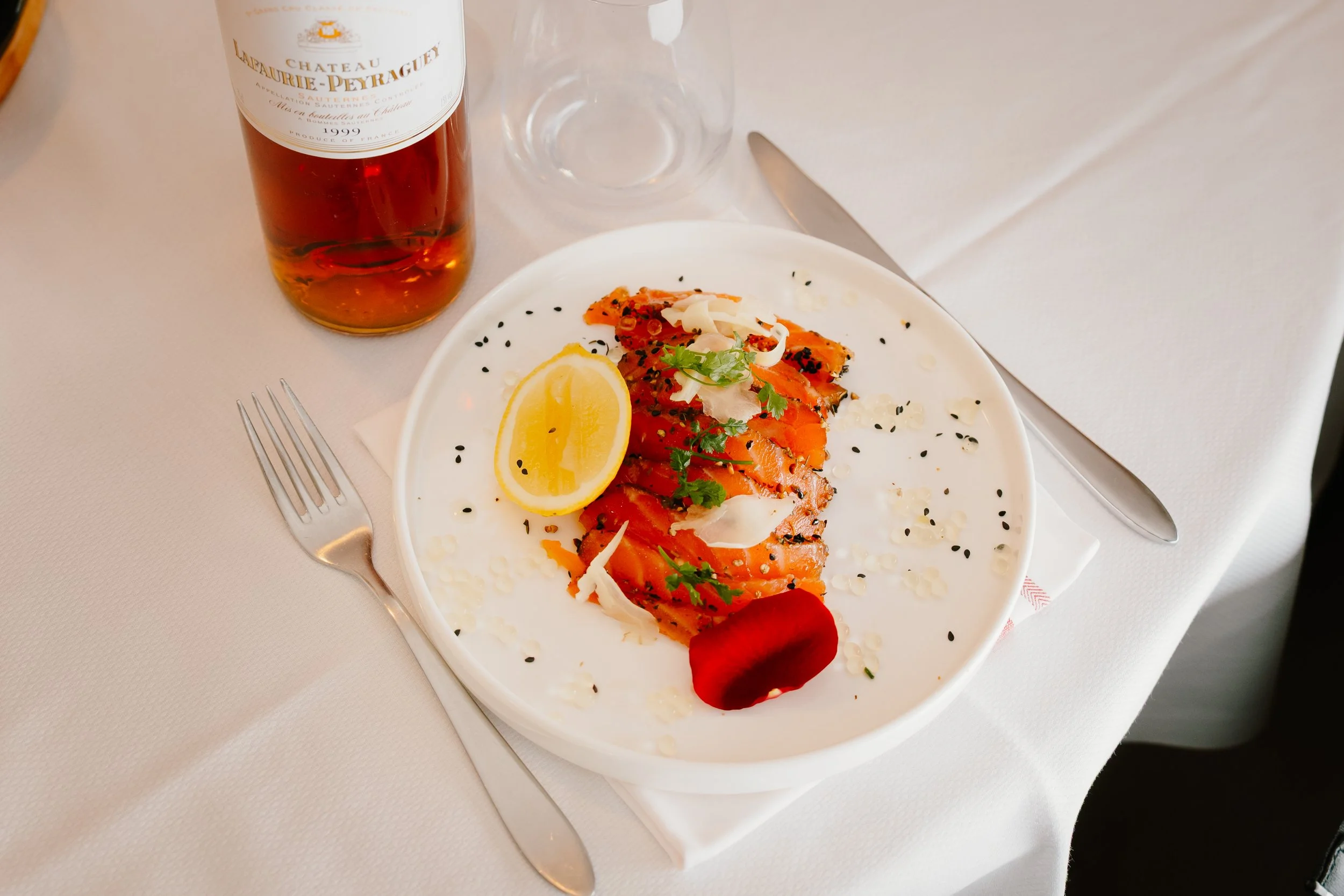 Chez Jaillet Lyon 6 | Assiette de saumon fumé avec une tranche de citron, garniture verte, graines de sésame noir et blanc, sur une table avec un verre vide, une bouteille de vin Château Laurencie-Peyraguey 1999 et des ustensiles.
