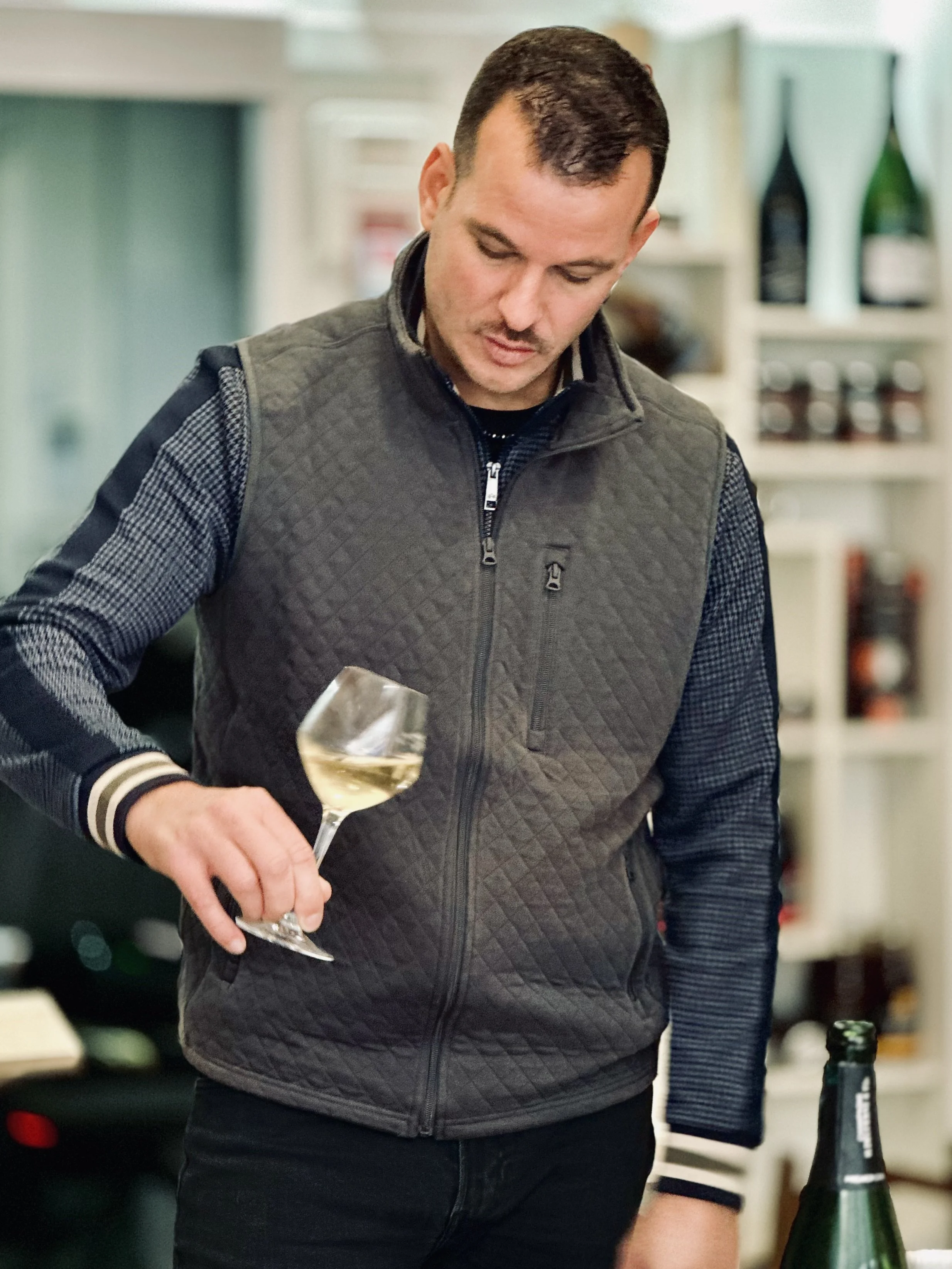 Chez Jaillet Lyon 6 | Alexis, le caviste de Chez Jailler, tient un verre de vin blanc dans une cuisine, avec des étagères contenant des bouteilles en arrière-plan. Il réalise une dégustation de vins dans sa cave à Lyon 6.