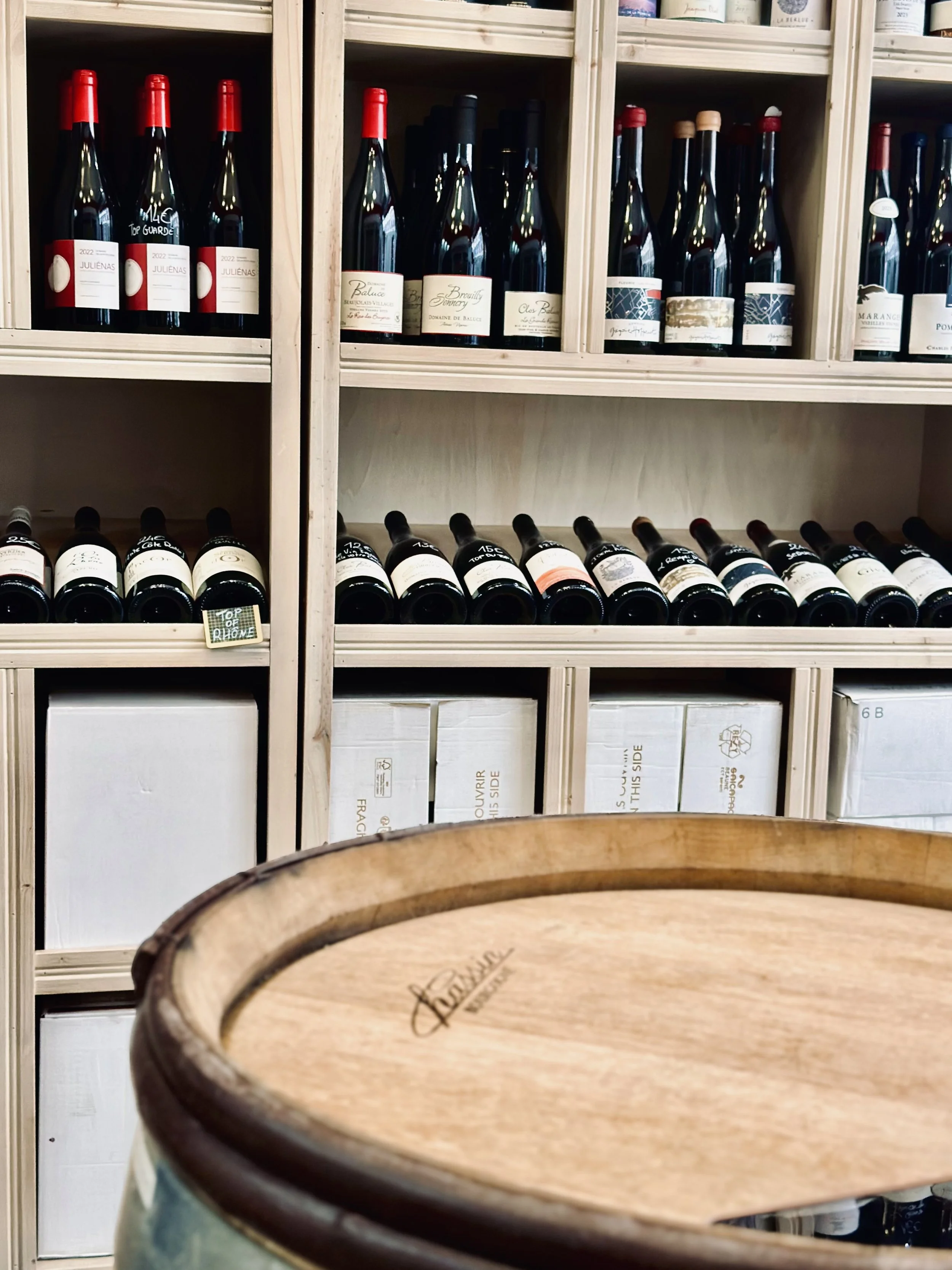 Chez Jaillet Lyon 6 | Une étagère en bois contenant plusieurs bouteilles de vin avec étiquettes colorées, et un tonneau en bois au premier plan dans la cave Chez Jaillet, Lyon 6.