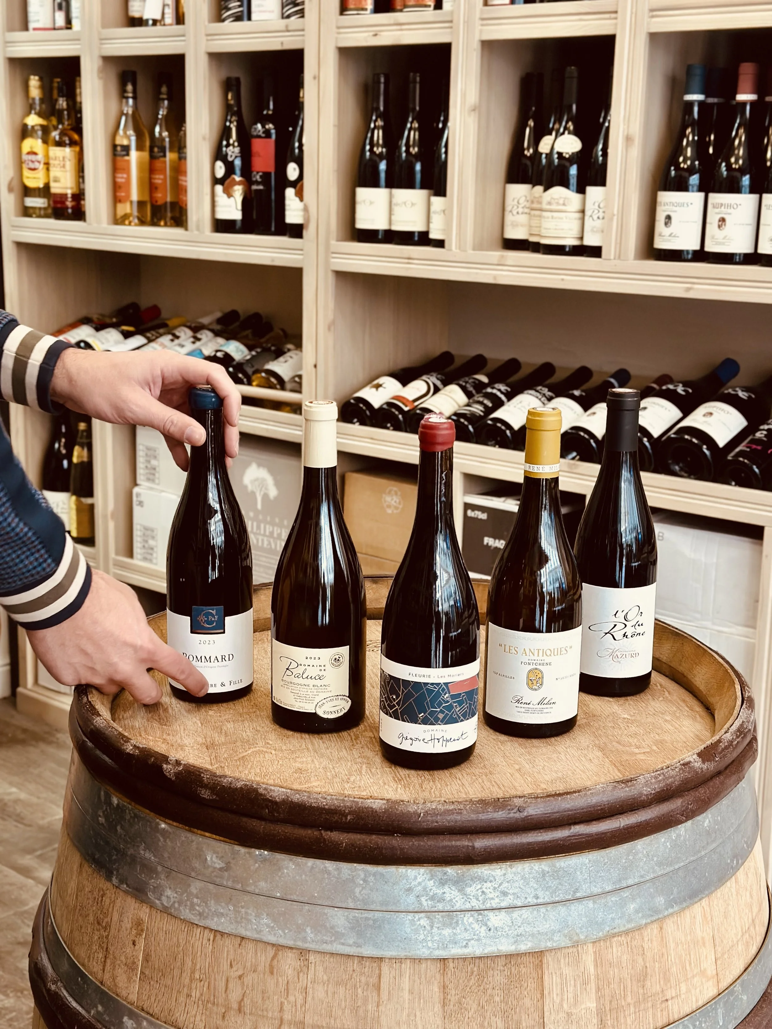 Chez Jaillet Lyon 6 | Cinq bouteilles de vin posées sur un tonneau en bois, avec des étagères de bouteilles de vin en arrière-plan dans une cave Chez Jaillet Lyon 6.