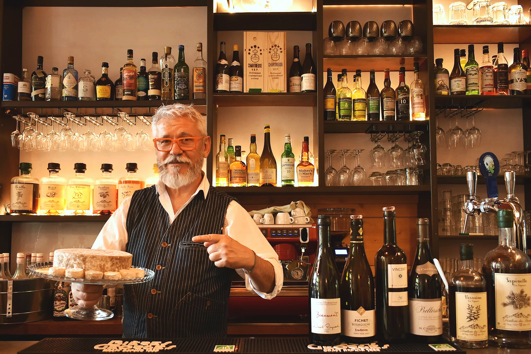Chez Jaillet Lyon 6 | Au bistrot, Didier Jaillet se tient derrière un bar, tenant un plateau de  fromages. Le fond montre des étagères avec des bouteilles d'alcool, des verres, et une machine à débit de boissons.