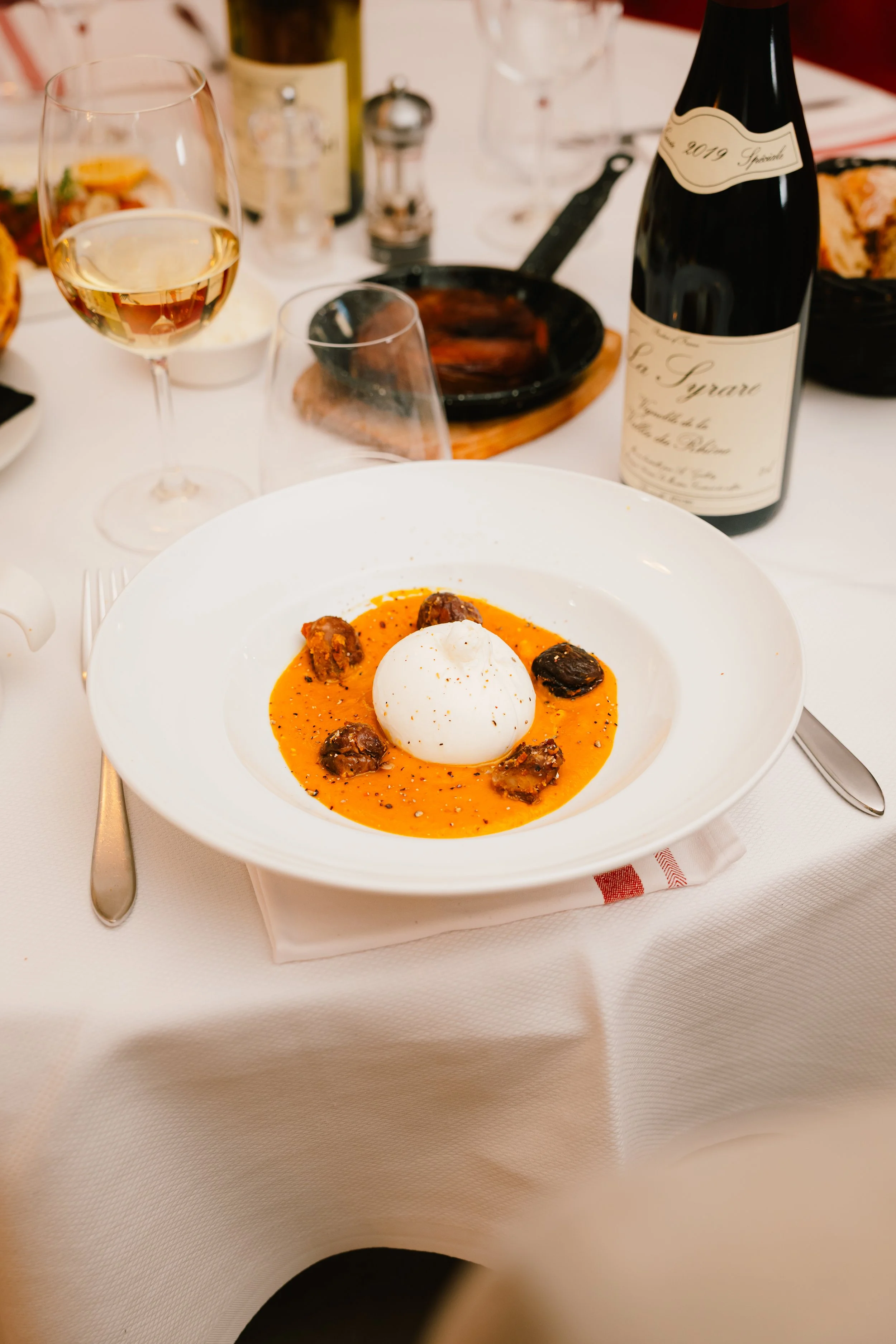 Chez Jaillet Lyon 6 | Assiette de mozzarella en sauce tomate avec morceaux de viande, sur une table de restaurant. Autour, une verre de vin blanc, une bouteille de vin rouge, et d'autres plats.
