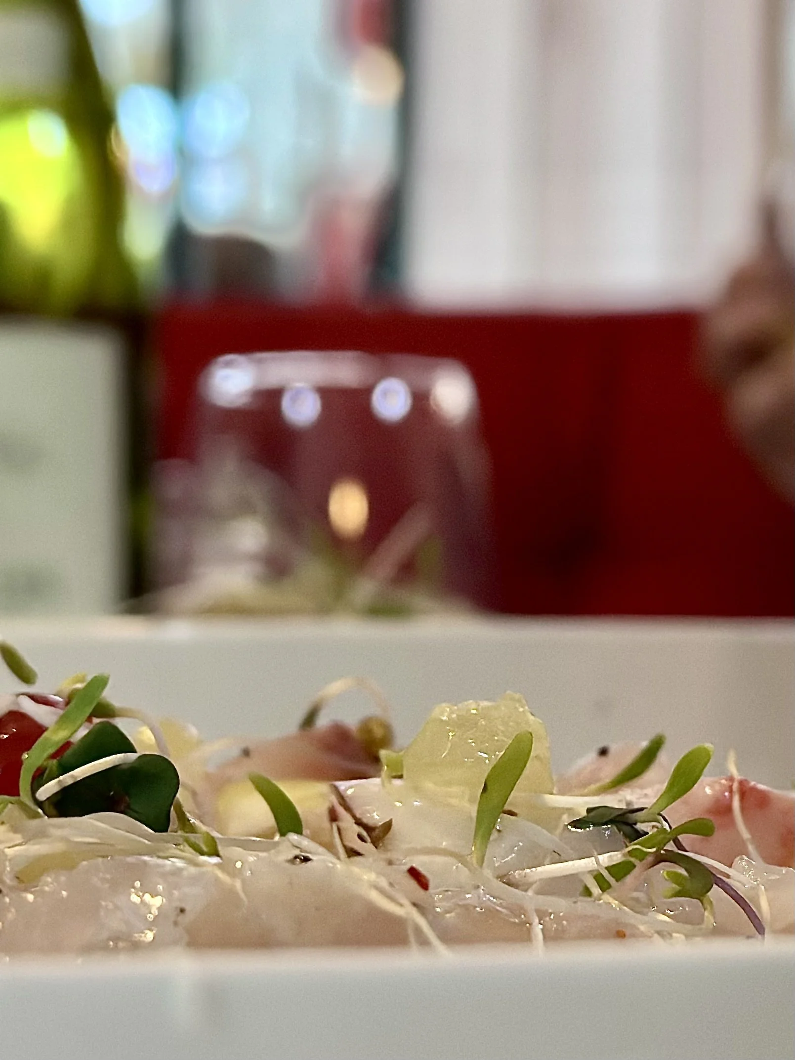 Chez Jaillet Lyon 6 | Une assiette avec des germes, des petits légumes et une sauce. En arrière-plan, une cuisine avec des verres et des meubles rouges, flous.