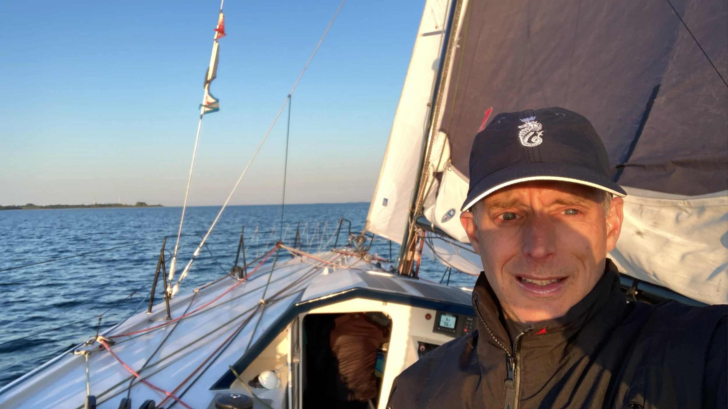 A man taking a selfie on a sailboat during sunset, with water and a distant shoreline in the background.