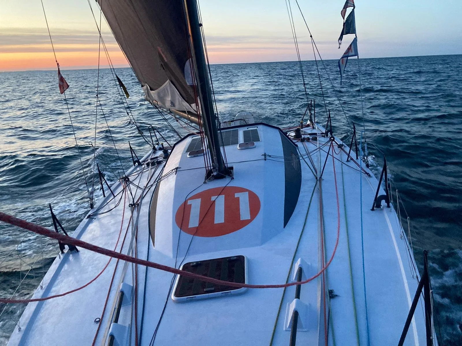 View from the deck of a sailboat at sunset with the ocean in the background, showing the boat's rigging, a large sail, and a red and white number 111 on the deck.