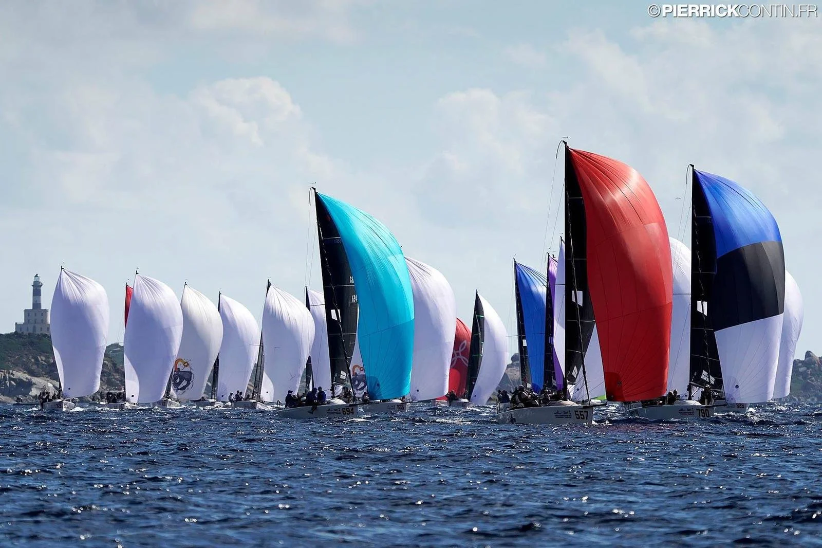 Multiple sailboats racing on open water, with colorful spinnakers of white, blue, red, and black, under a partly cloudy sky. A lighthouse is visible on a distant hill in the background.