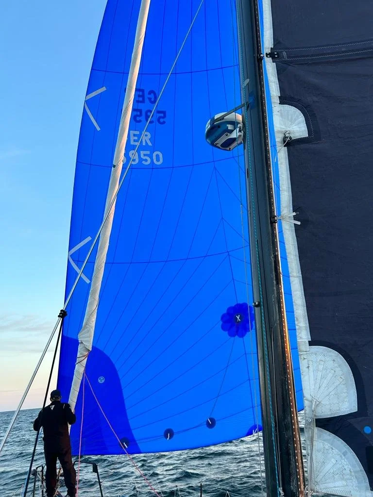 A person on a sailboat steering the boat on the water, with a large blue sail and part of the boat's structure visible.