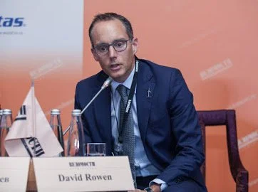 A man in a dark suit, glasses, and a tie seated at a conference table with nameplate 'David Rowen,' speaking into a microphone.