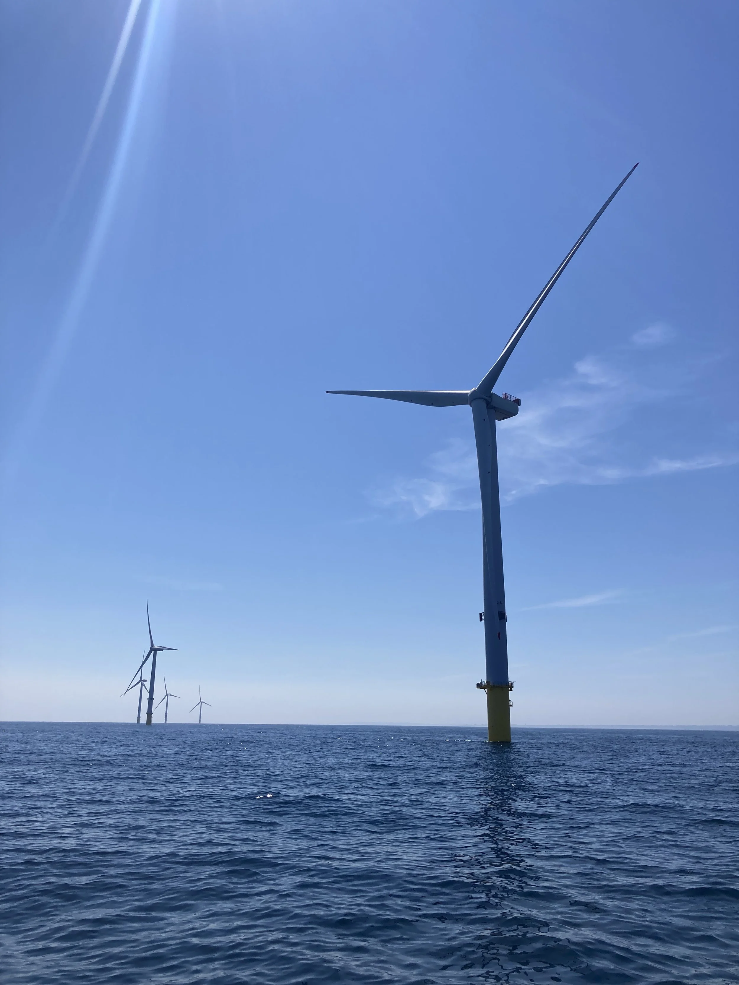 Several offshore wind turbines in the ocean under a clear blue sky.