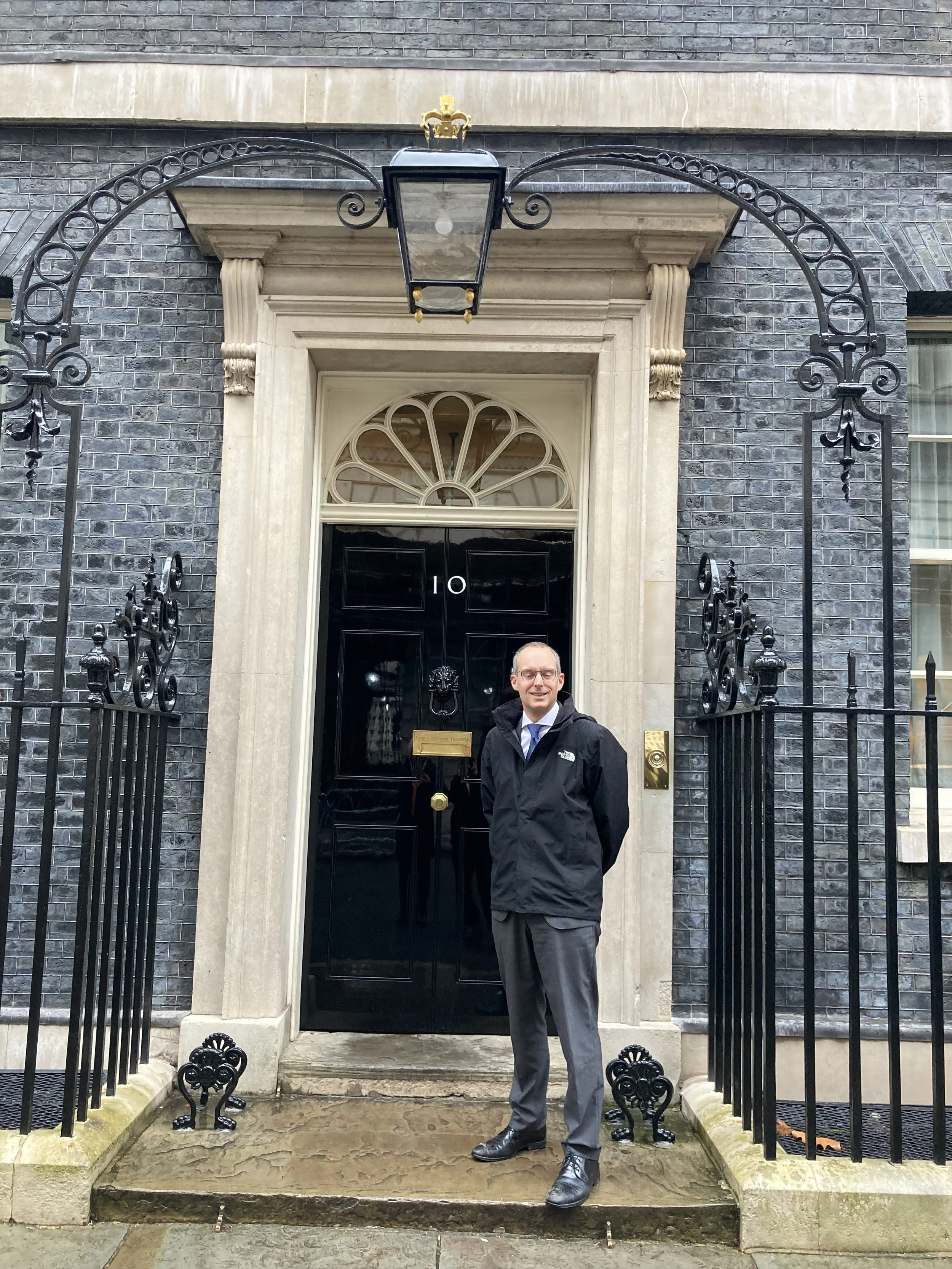 A man in a black jacket and gray trousers standing in front of a black door with the number 10. The door is framed by a white stone surround and topped with a fanlight window. The building exterior is dark gray brick with ornate iron fencing and a lantern above the door.