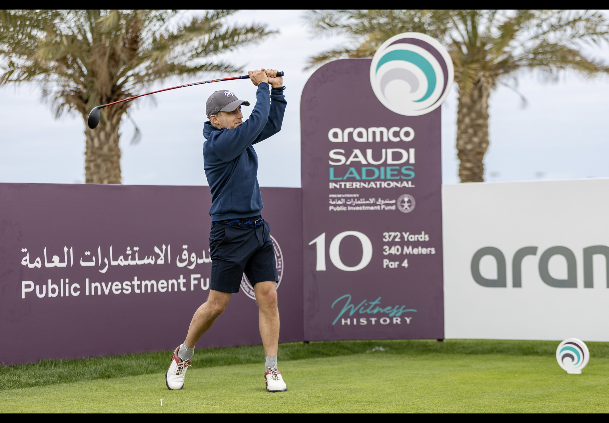 A man playing golf on a course with a purple sign behind him for the Aramco Saudi Ladies International golf tournament, showing details about the 10th hole, which is 372 yards and a par 4, with palm trees and cloudy sky in the background.