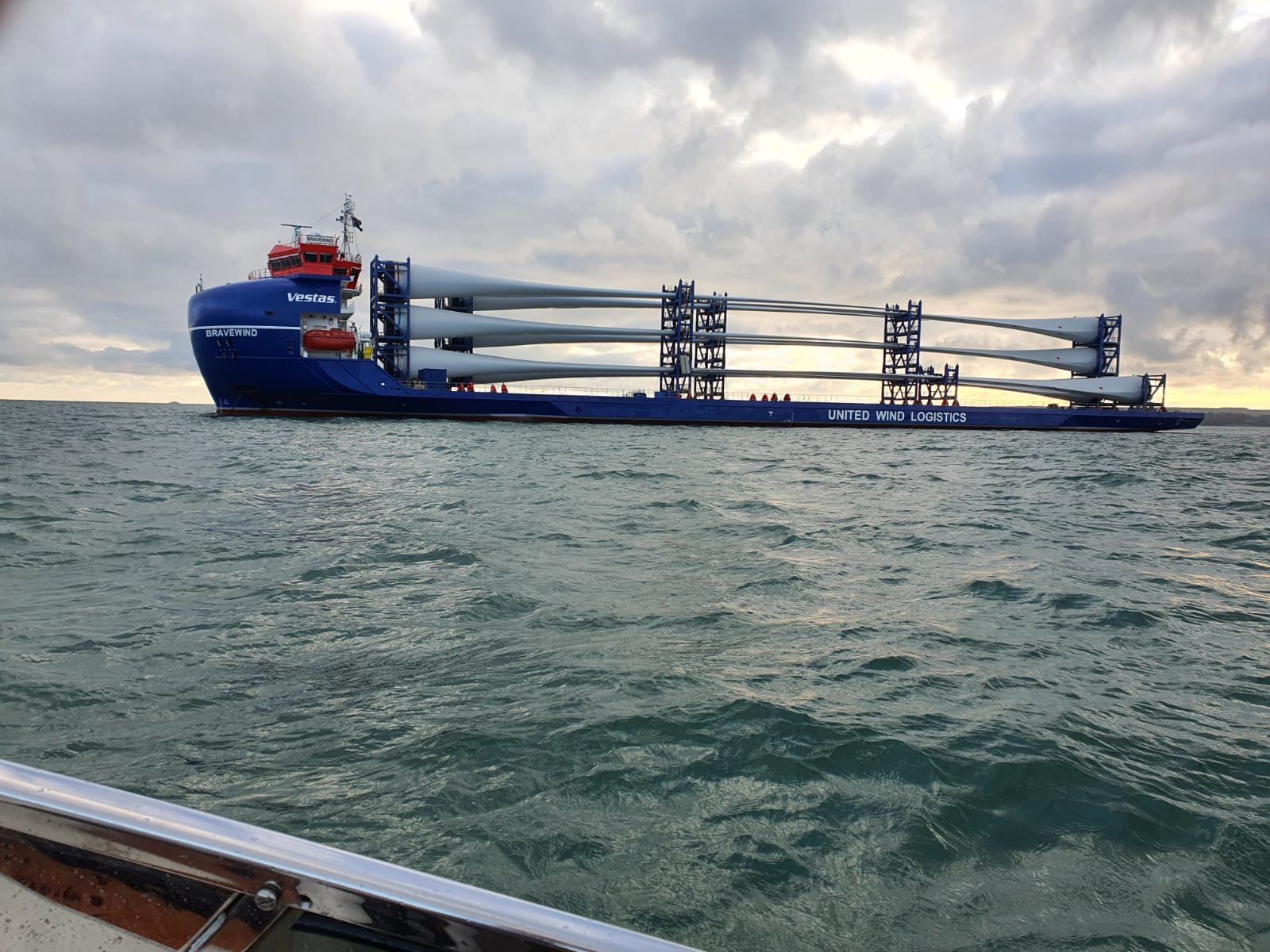 A large blue wind turbine installation vessel named 'Bravewind' on the water, with the words 'United Wind Logistics' on its side, under cloudy sky.