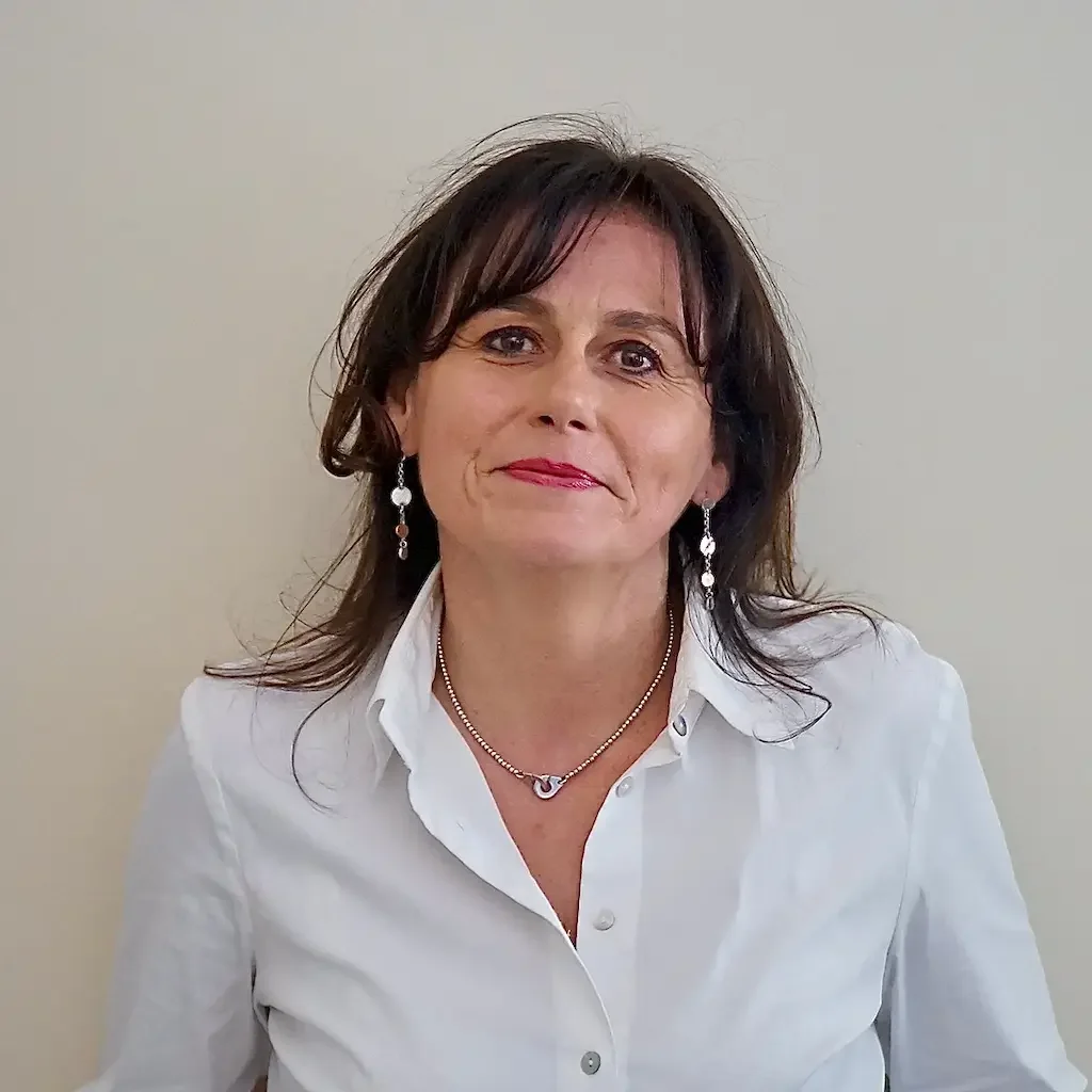Une femme souriante avec des cheveux bruns, portant un chemisier blanc, des boucles d'oreilles pendantes et un collier, posant devant un mur clair.