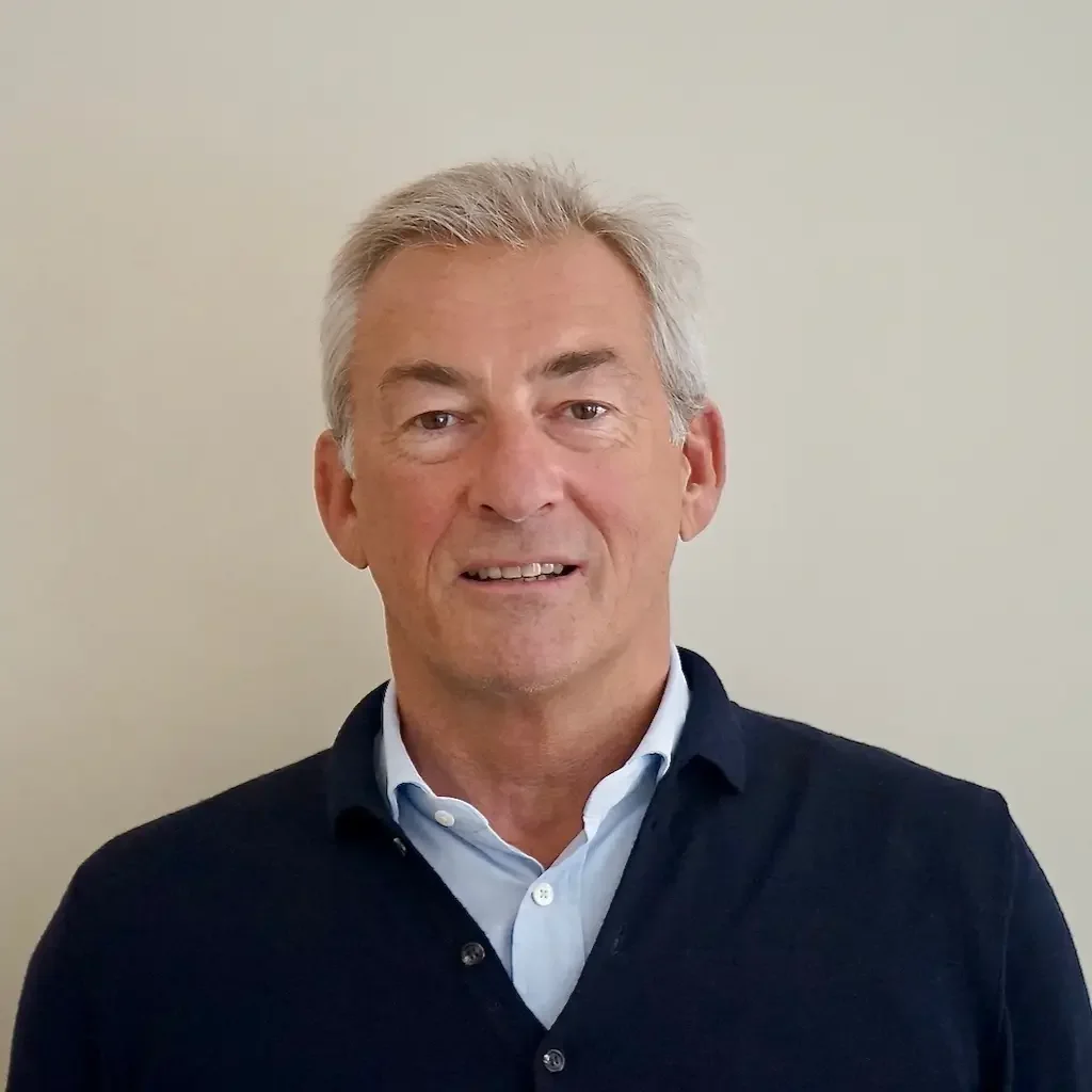 Un homme d'âge moyen avec des cheveux gris, portant une chemise blanche et un pull sombre, pose devant un mur beige clair.