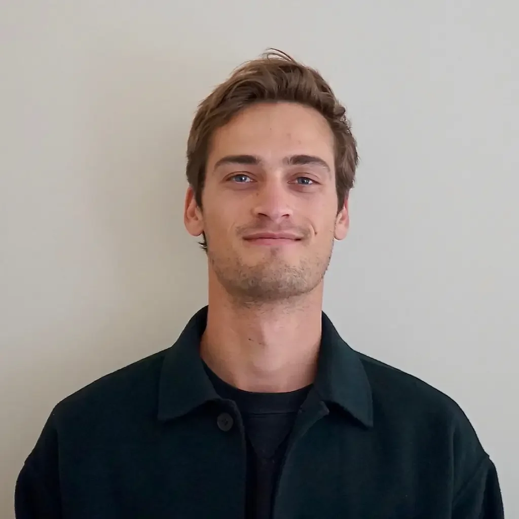 Jeune homme avec cheveux bruns, portant une veste noire, souriant légèrement, devant un mur blanc.