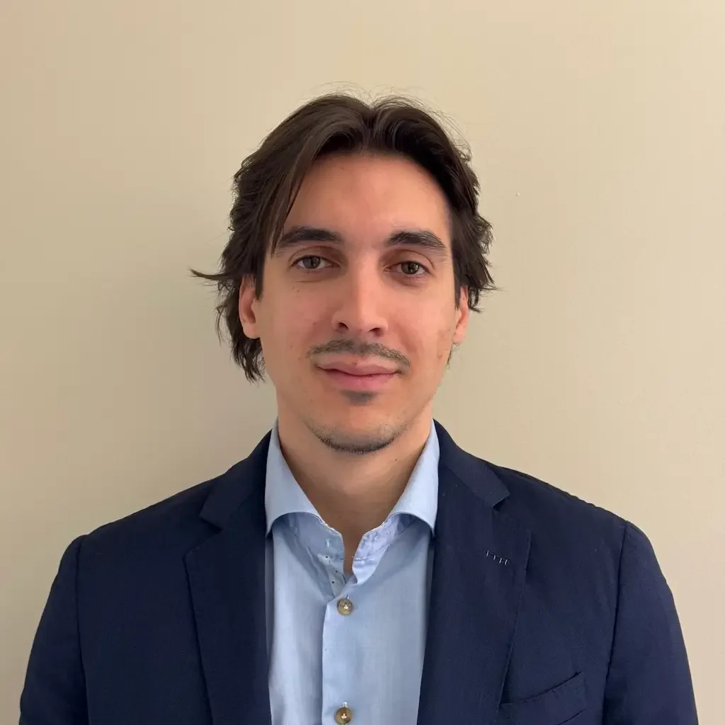 Portrait d'un jeune homme avec des cheveux bruns, portant une chemise bleue et un blazer noir, devant un mur beige clair.