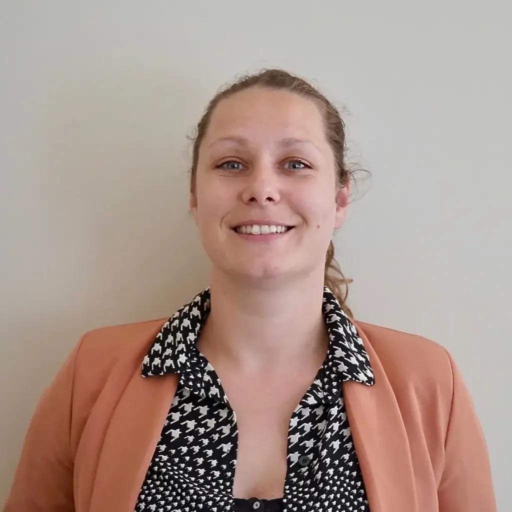 Une jeune femme souriante avec des cheveux bouclés attachés, portant un blazer orange et une chemise à motifs en noir et blanc, pose devant un mur beige.