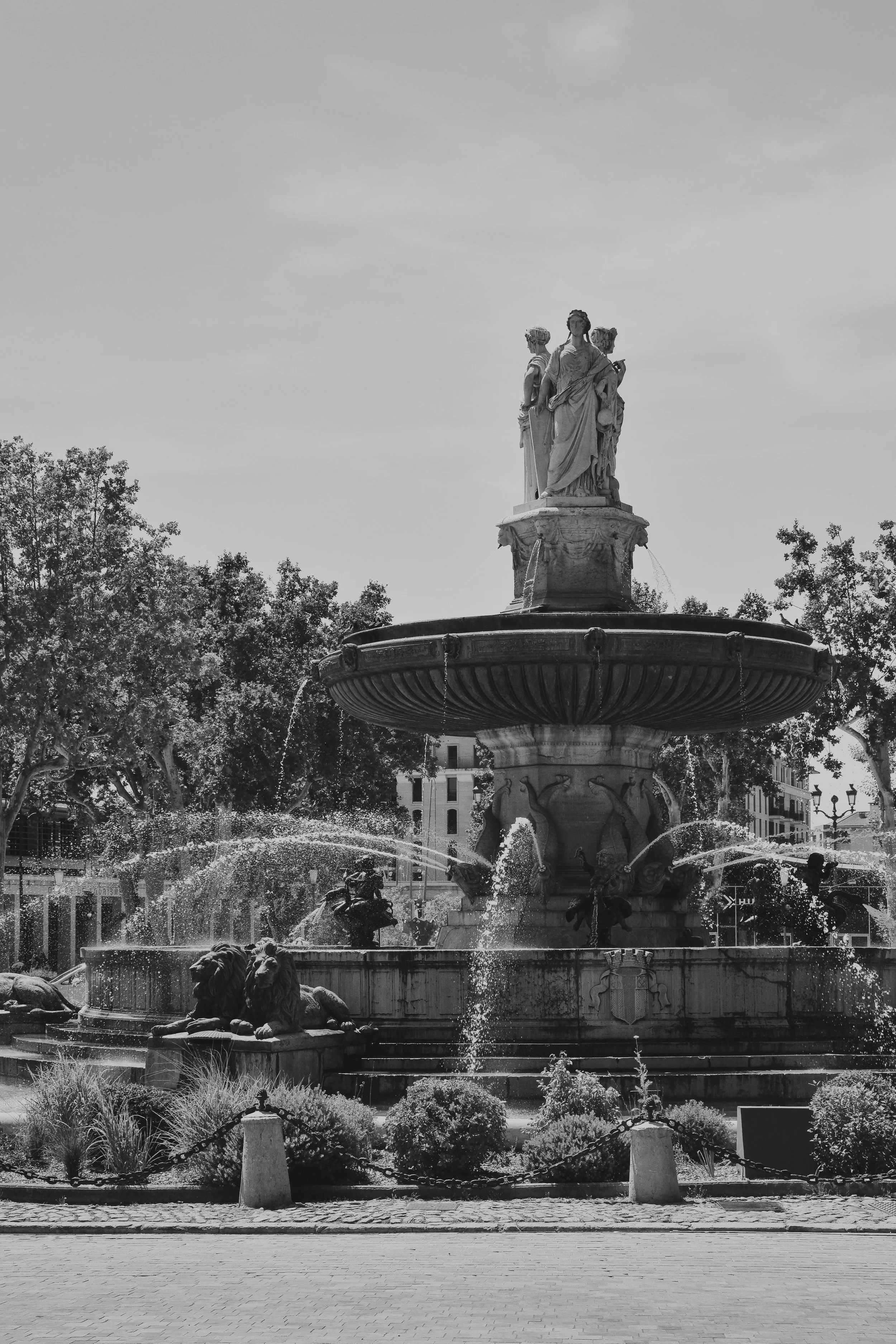 Une fontaine ornée de sculptures de figures féminines et de lions en bas, entourée de végétation, dans une ville, en noir et blanc.