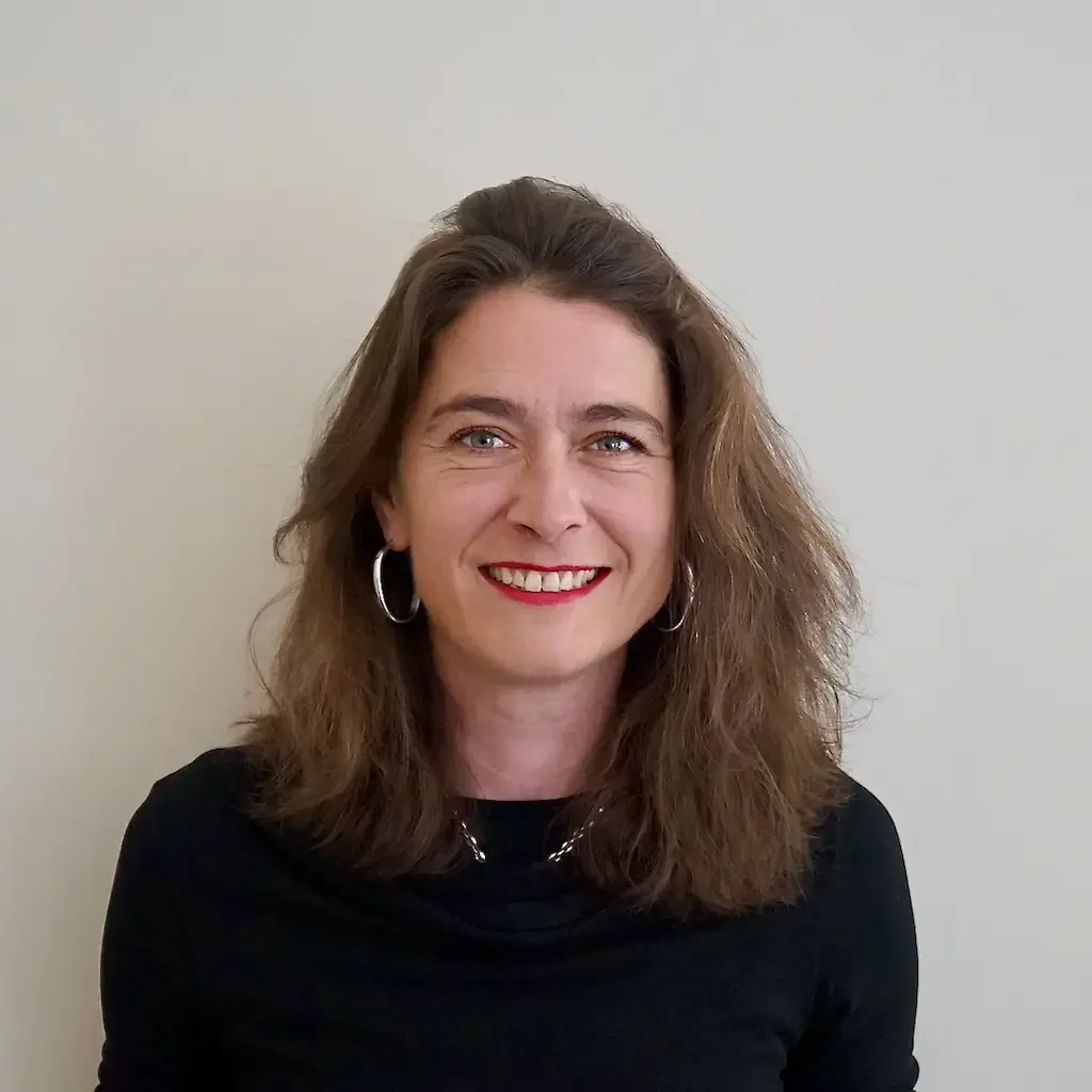 Une femme aux cheveux bruns bouclés, portant des boucles d'oreilles en anneau et un chandail noir, souriante devant un mur beige.