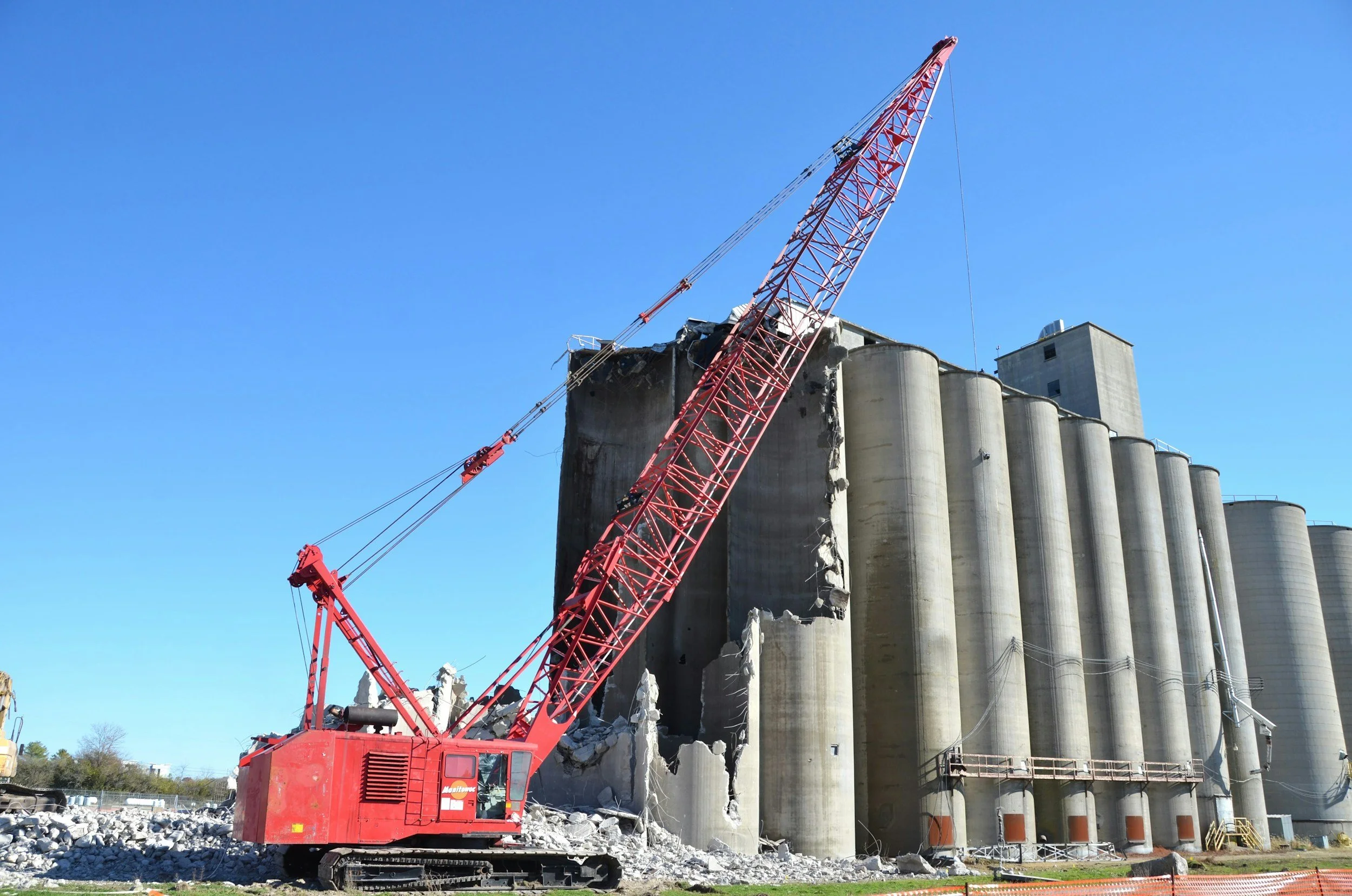 A large red crane demolishing a concrete industrial structure with cylindrical silos under a clear blue sky.