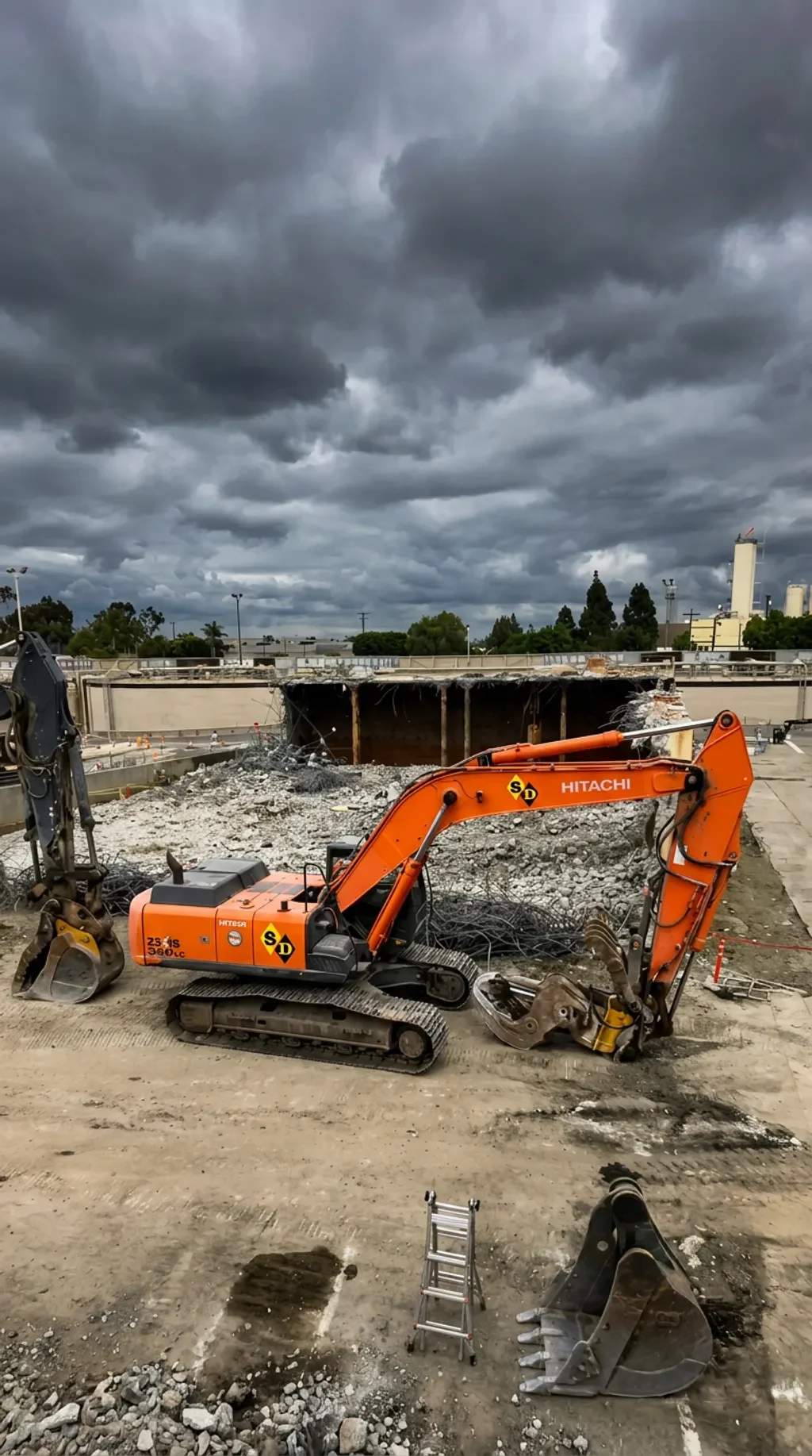 Construction site with a small orange excavator, piles of debris, a large hole in the ground, and dark stormy clouds overhead.