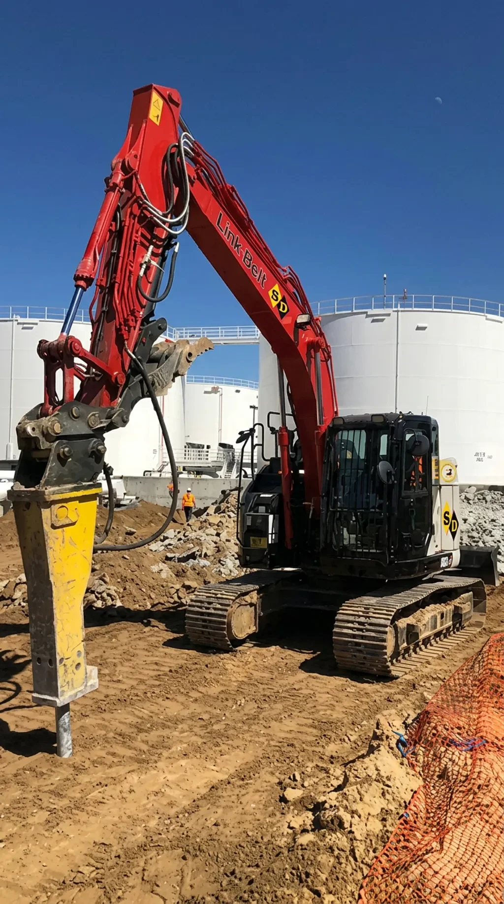 A red excavator with tracked wheels is working on a construction site, digging into the dirt near large white storage tanks and underground pipes. There are three workers in orange safety vests and helmets in the background.