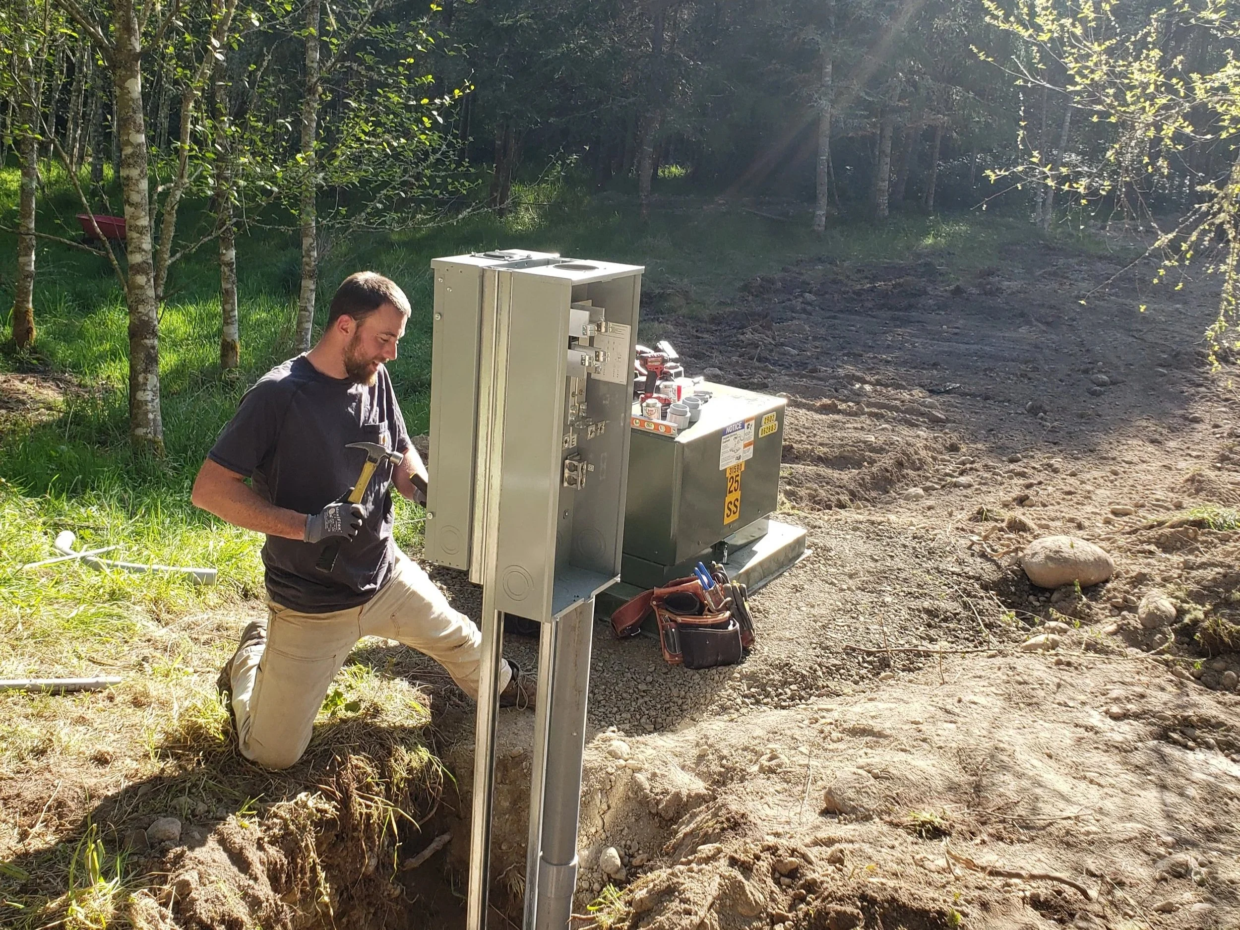 A man kneeling on the ground working on an electrical box in a wooded outdoor setting during daytime.